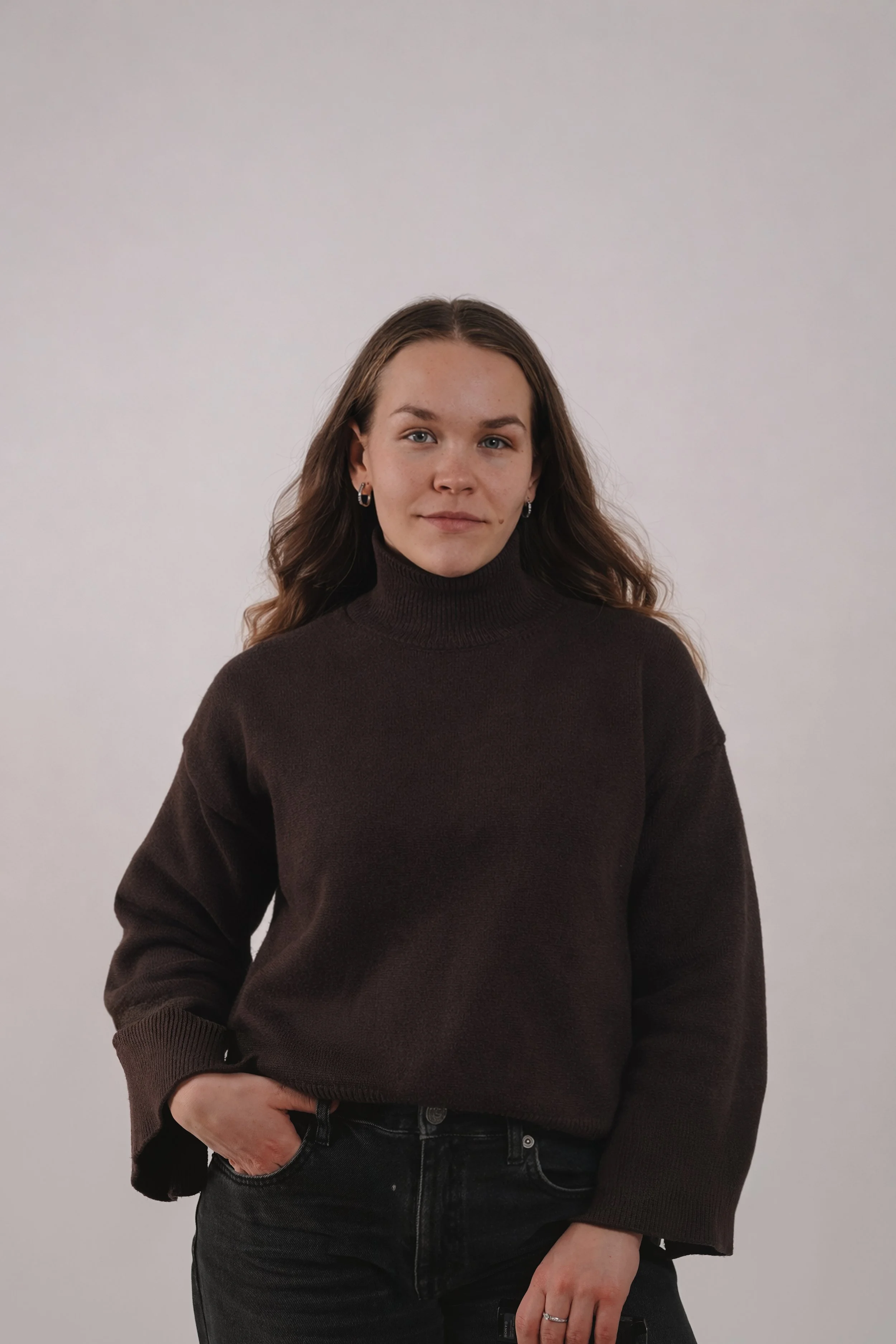 Junge Frau mit braunem Pullover, schwarzer Jeans, langen braunen Haaren und Ohrringen vor weißem Hintergrund.