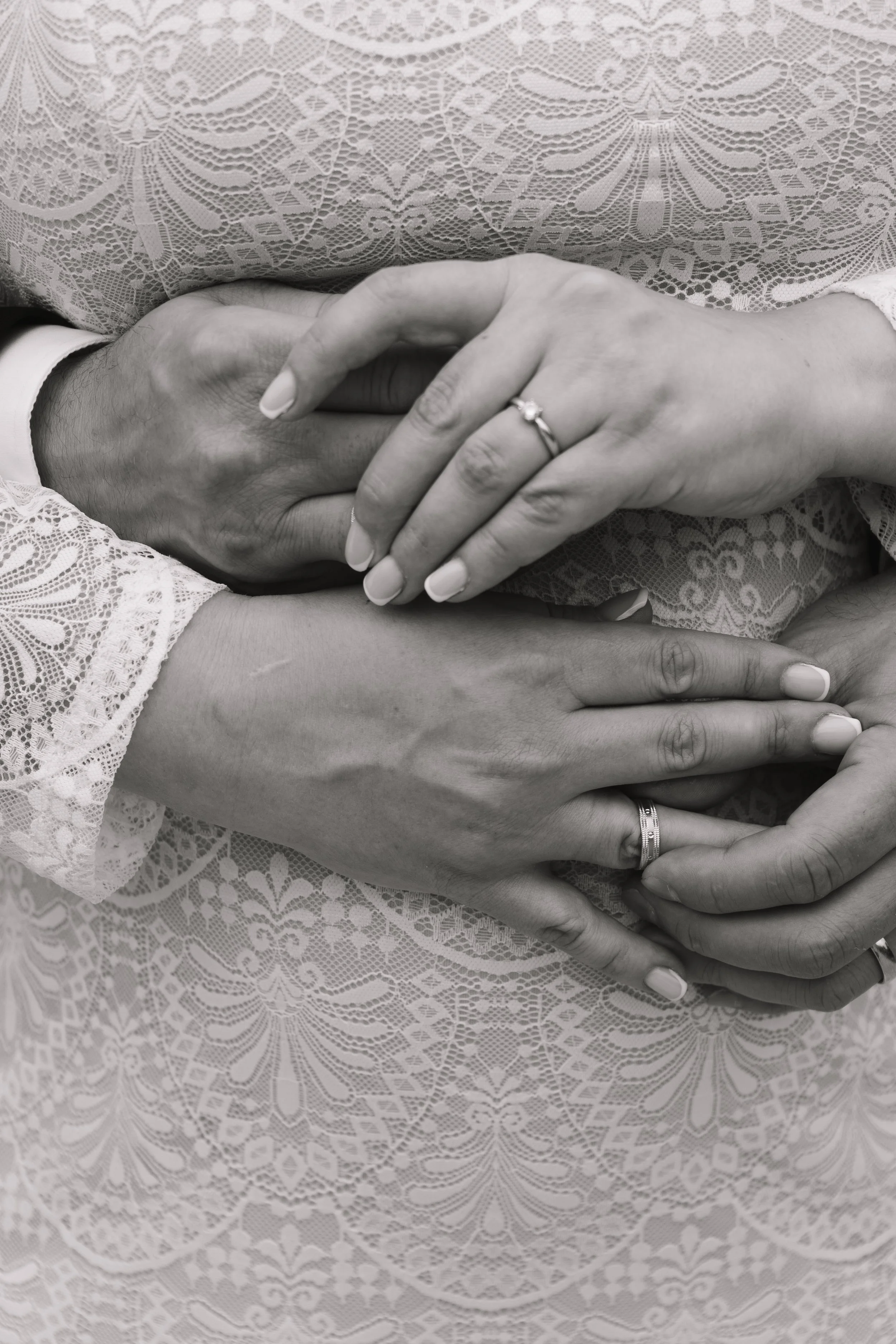 Schwarz-weiße Nahaufnahme von Händen, die sich in einer symbolischen Geste halten, mit Eheringen, auf einem eleganten, Spitzenstoff-Kleid