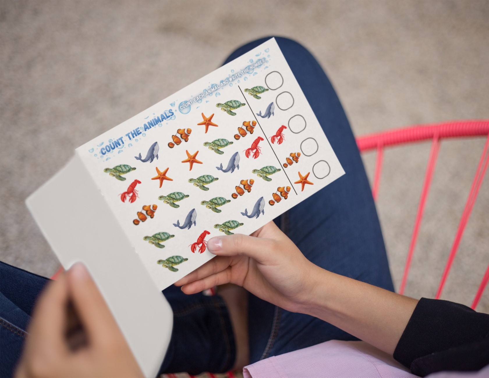 A person holding a sheet of paper with various sea animals and counting circles, sitting on a pink chair.