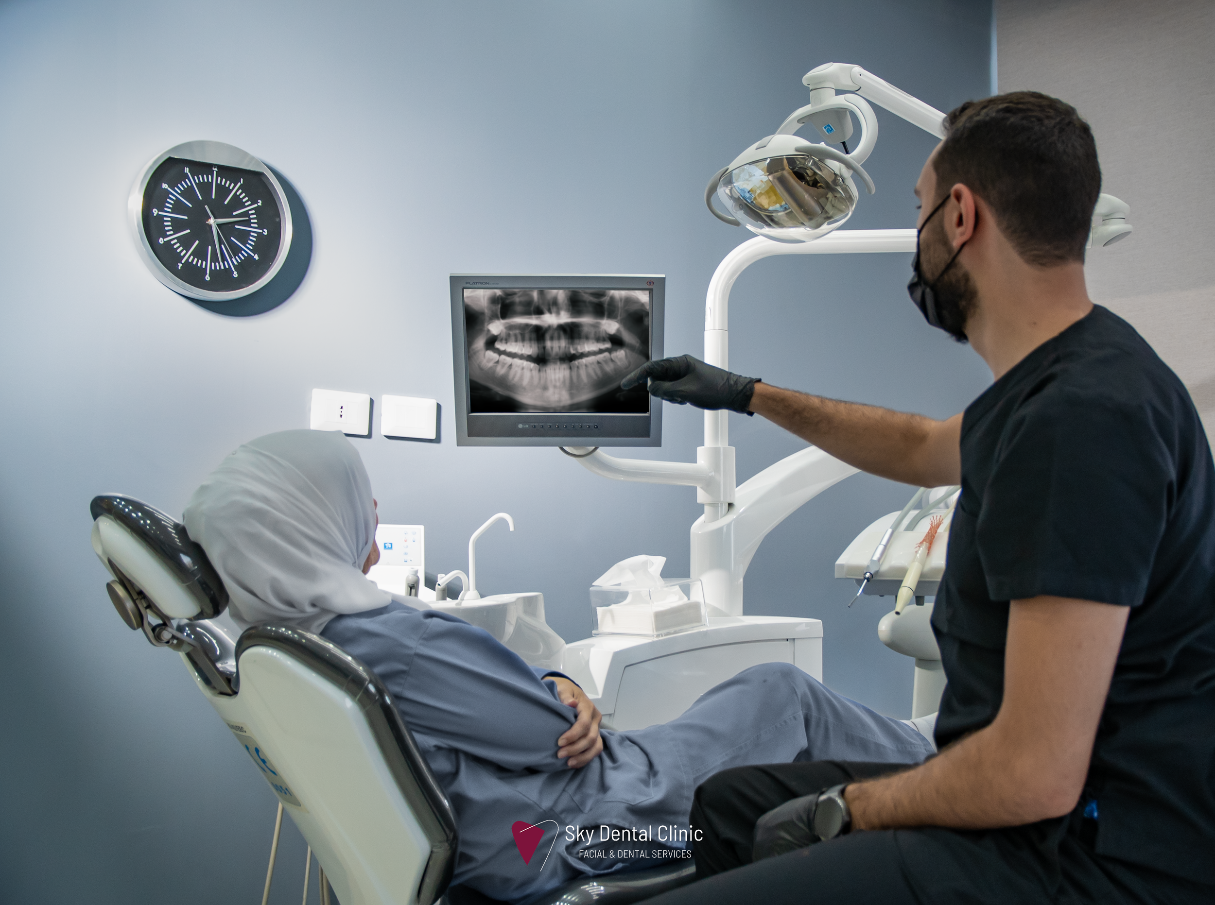 A dentist and a patient in a dental clinic. The dentist is pointing at an X-ray on a monitor while sitting on a stool. The patient is seated in a dental chair, wearing a head cover. Dental equipment and a clock are visible on the blue and white walls.