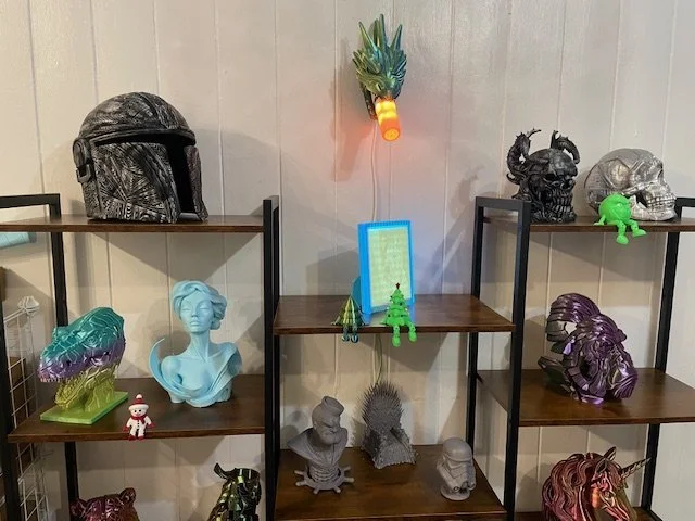 Decorative shelves with eclectic items including a Mandalorian helmet, a pineapple wall light, a green Rilakkuma figure, a bust of a woman, a small Santa figurine, artistic skull sculptures, a creature resembling a starfish, and colorful busts, all arranged on dark wooden and metal shelving units.