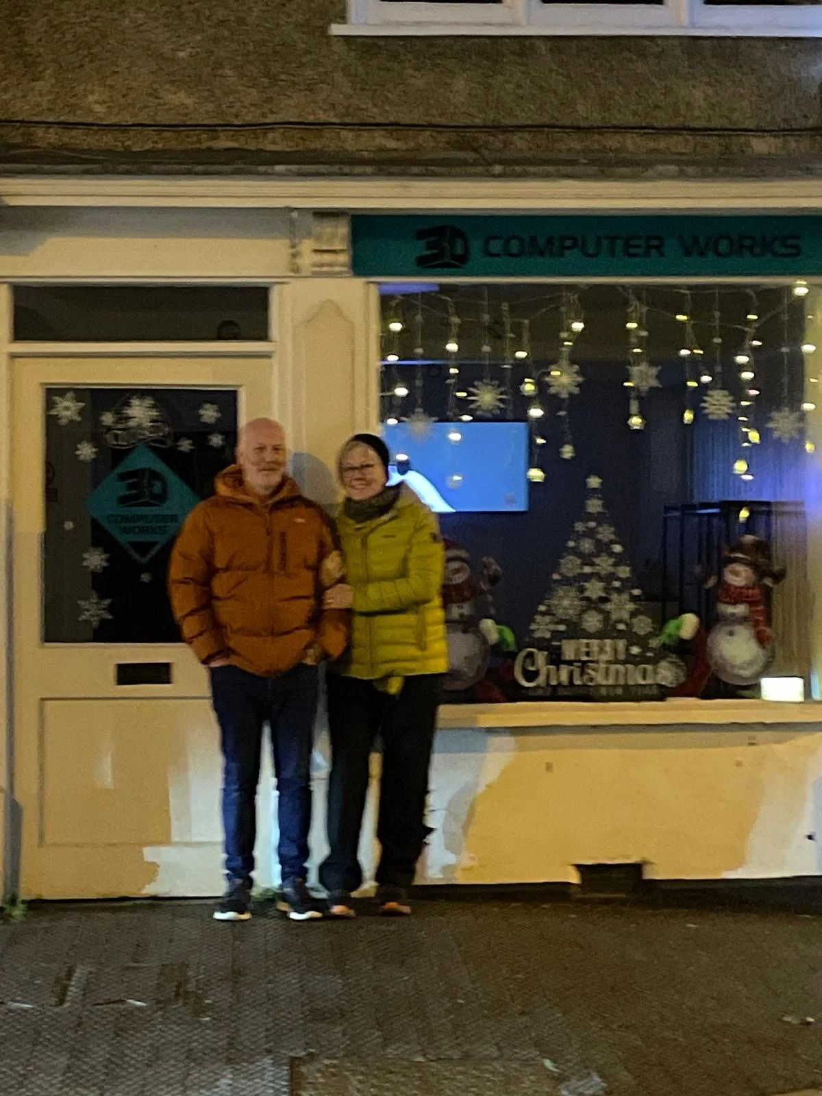 Two people standing outside a holiday-decorated shop, smiling, wearing winter jackets.