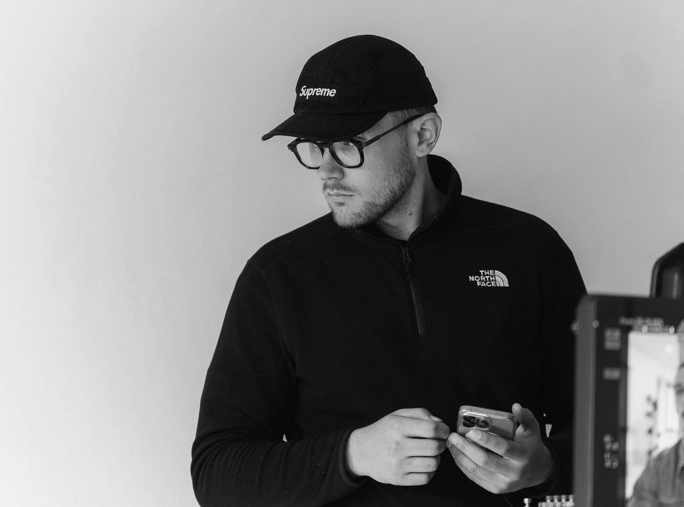 A man wearing glasses, a black cap with 'Supreme' logo, and a black North Face jacket, looking at his phone against a plain wall.