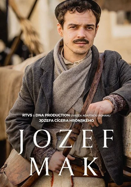 A man dressed in period clothing, with a mustache, sitting outdoors with a serious expression, holding a rifle, with a rustic background.
