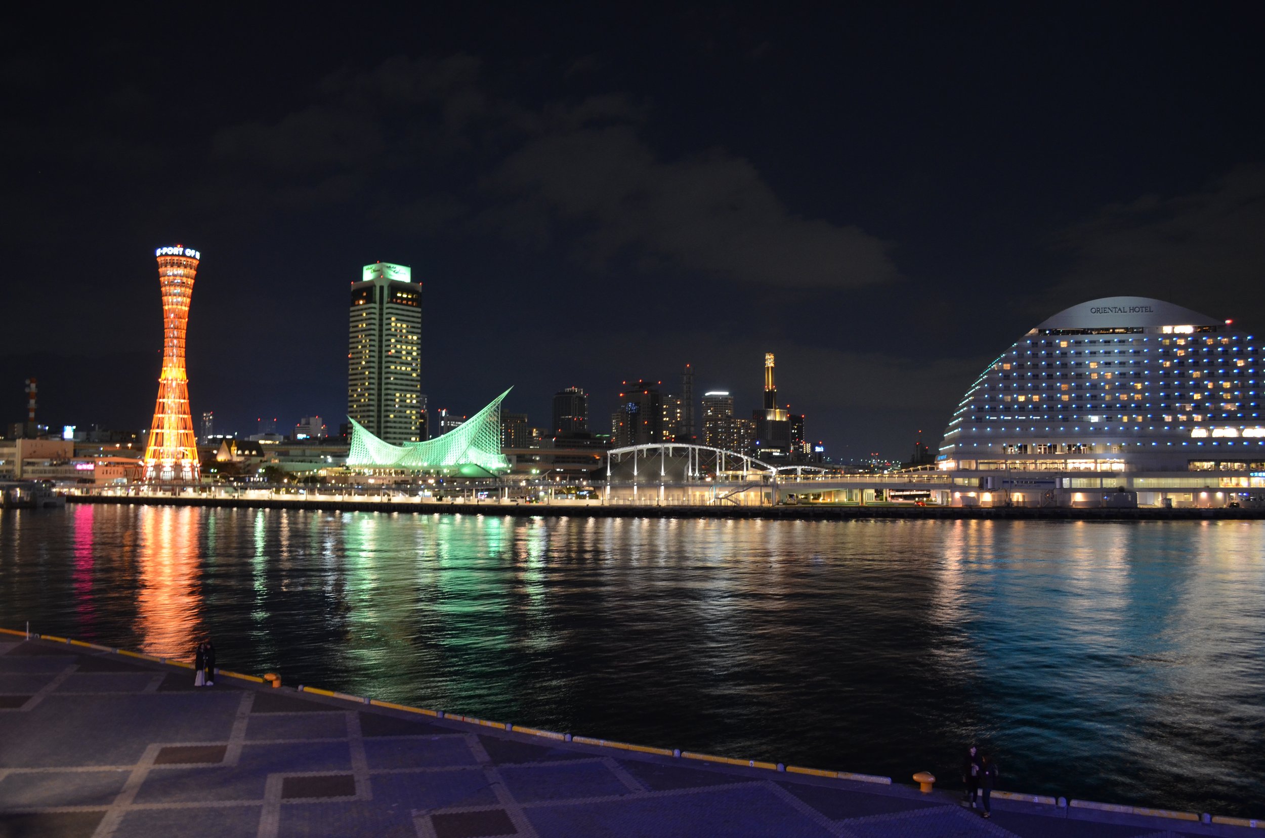 Vista nocturna del skyline del puerto con edificios iluminados reflejándose en el agua, incluyendo una torre naranja, un edificio alto con luces verdes, y un hotel grande de cúpula iluminada.
