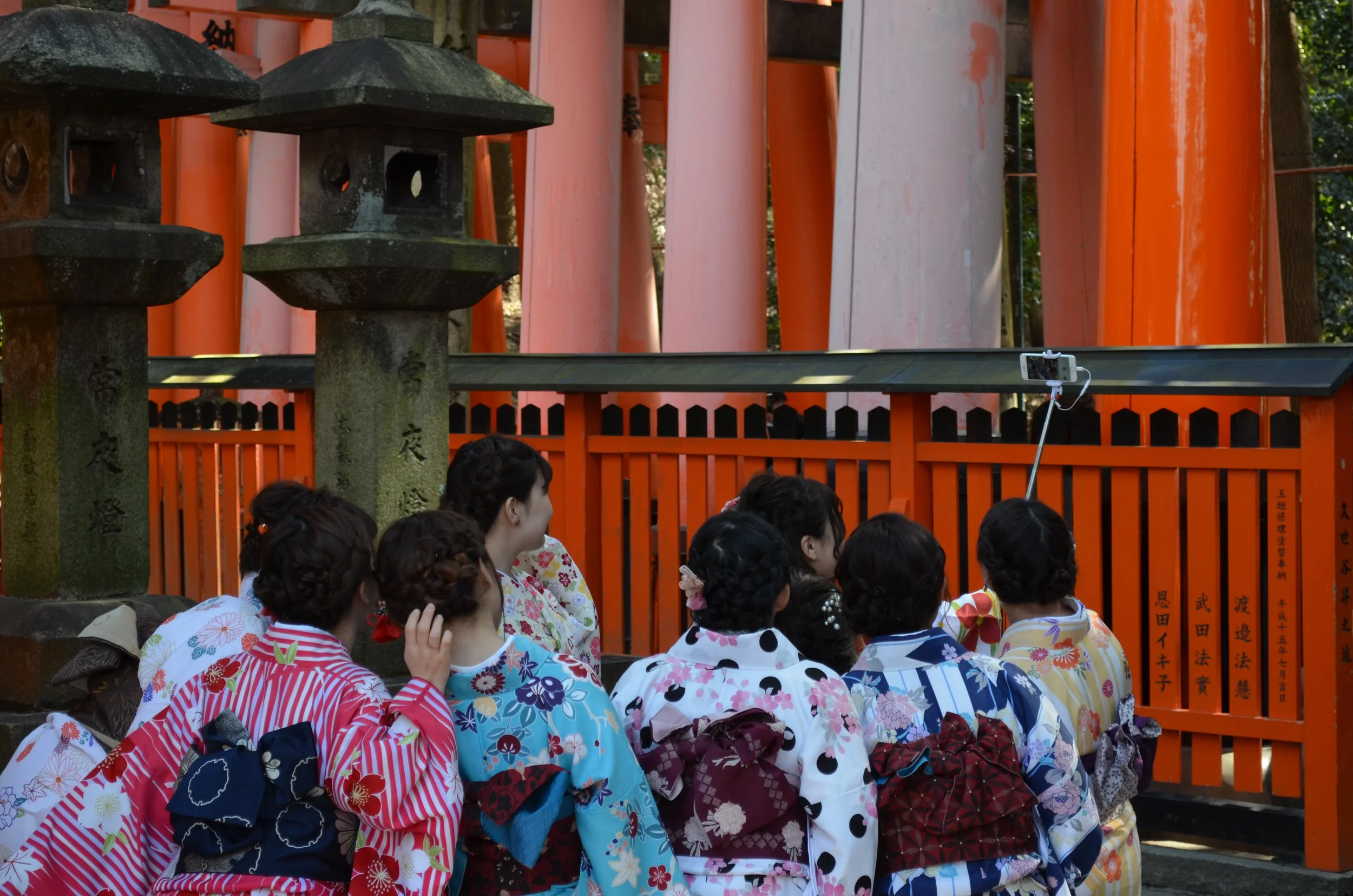 Grupo de mujeres jóvenes vestidas con kimonos tradicionales japoneses tomando una selfie frente a un santuario con linternas de piedra y puertas torii de color naranja.