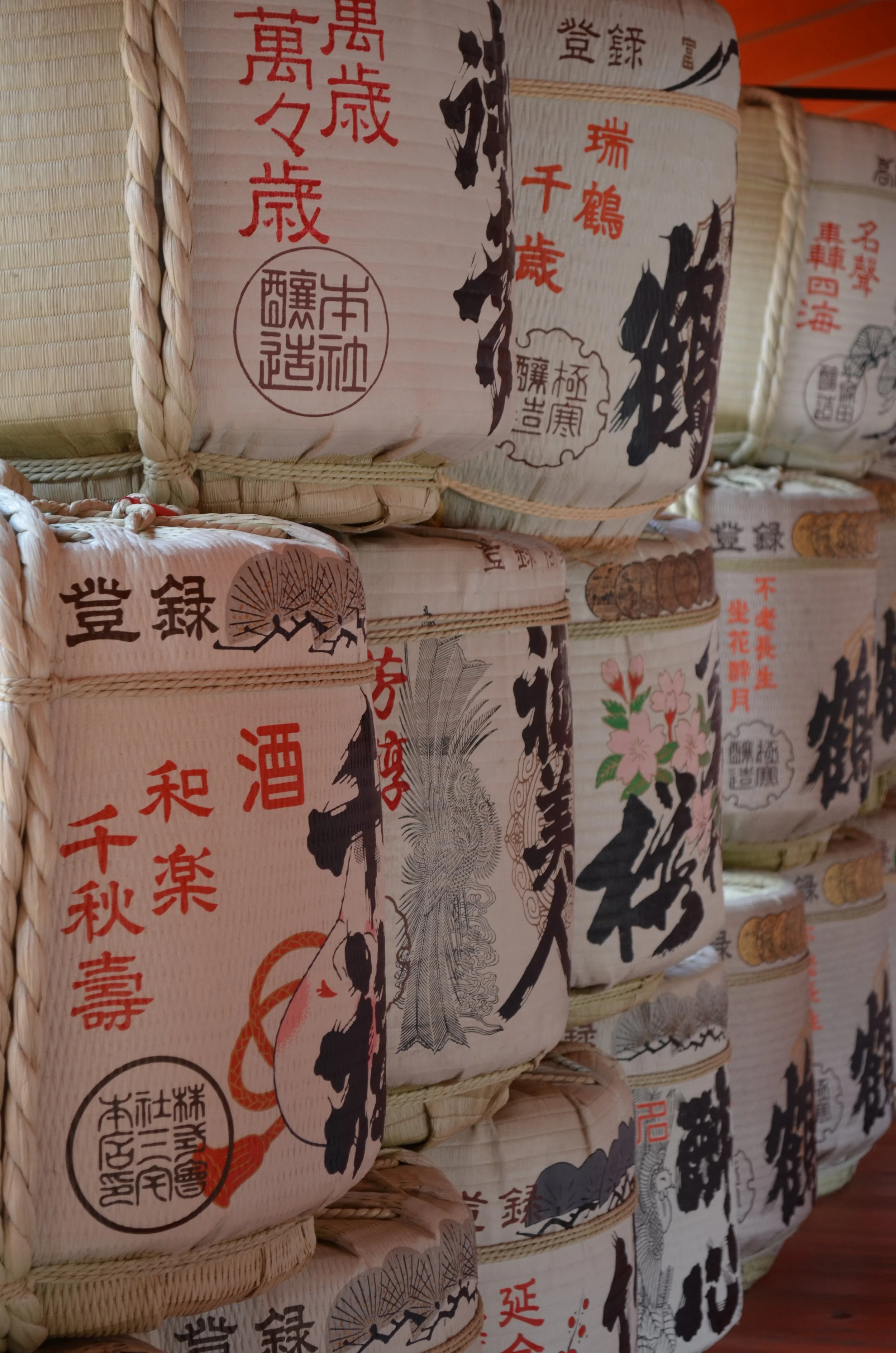 Varios barriles tradicionales de sake japoneses apilados, decorados con etiquetas con caracteres kanji y ilustraciones de flores, montañas y paisajes.