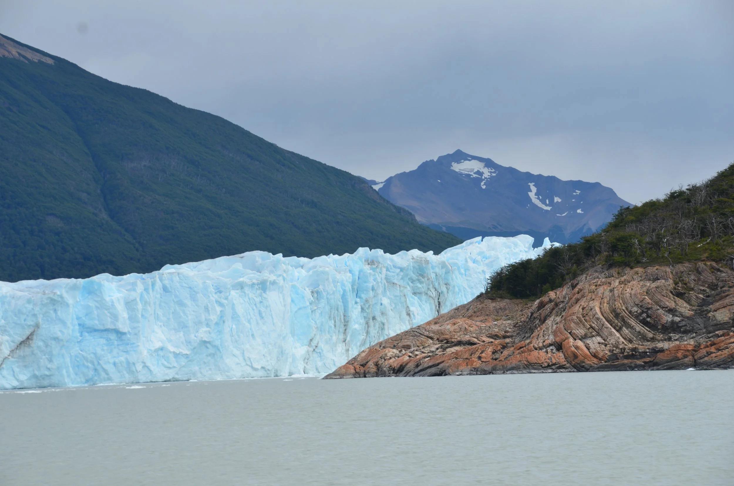 Glaciar azul en un paisaje montañoso, con agua en primer plano, bosques y picos nevados en el fondo.