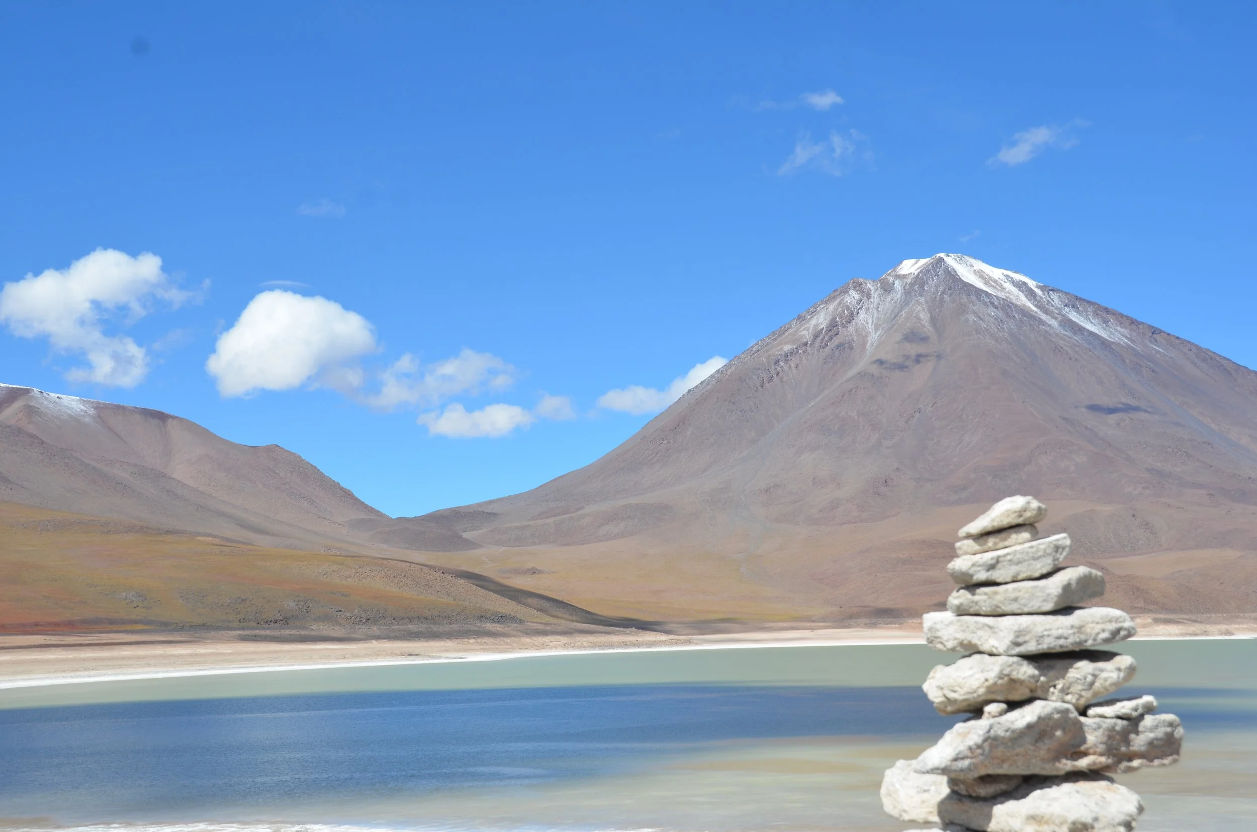 Montañas con nieve y un lago en primer plano, con un montón de piedras apiladas en la orilla.
