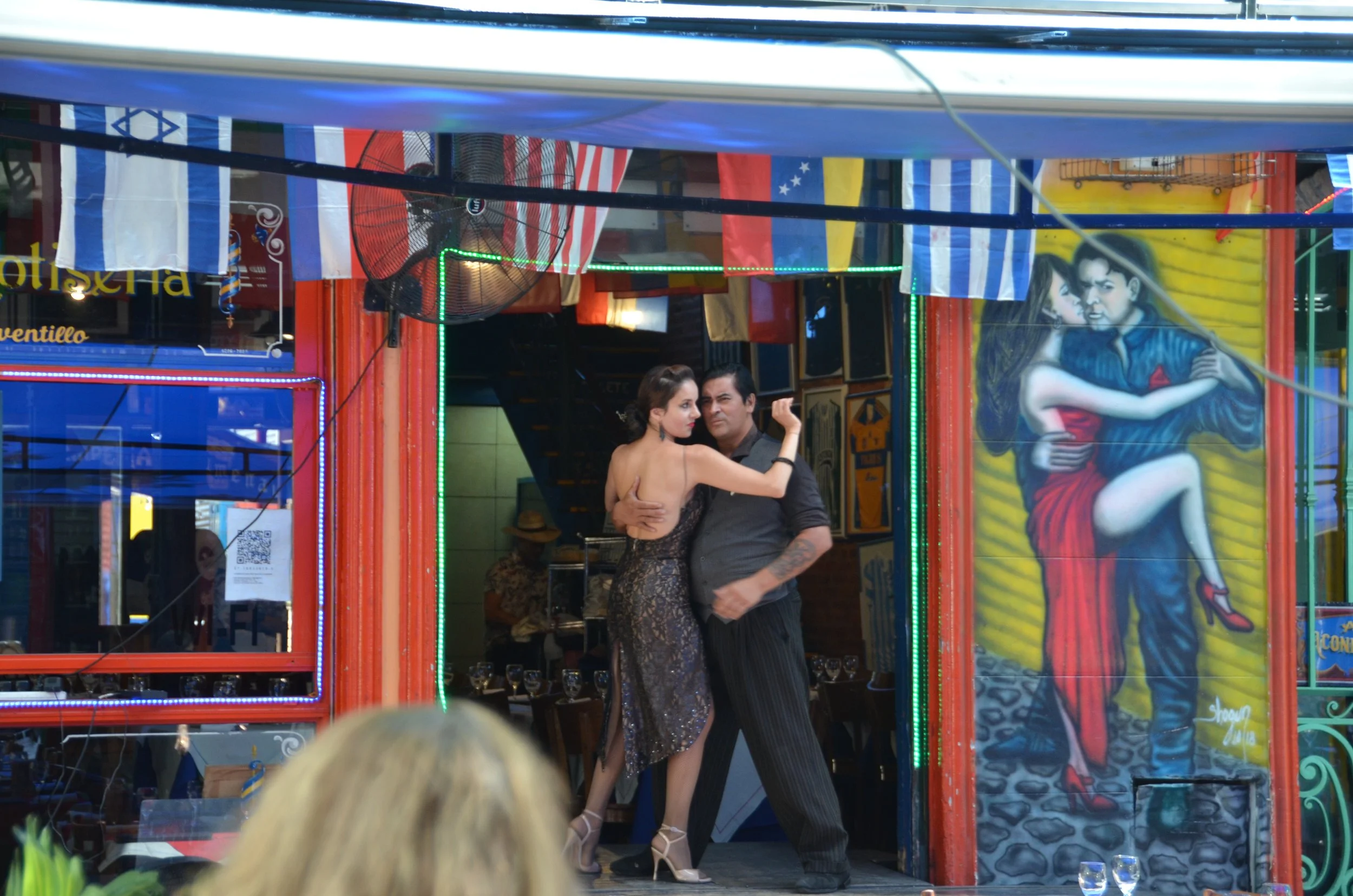 Dos personas bailando tango en un restaurante decorado con banderas de varios países y arte mural en la pared.