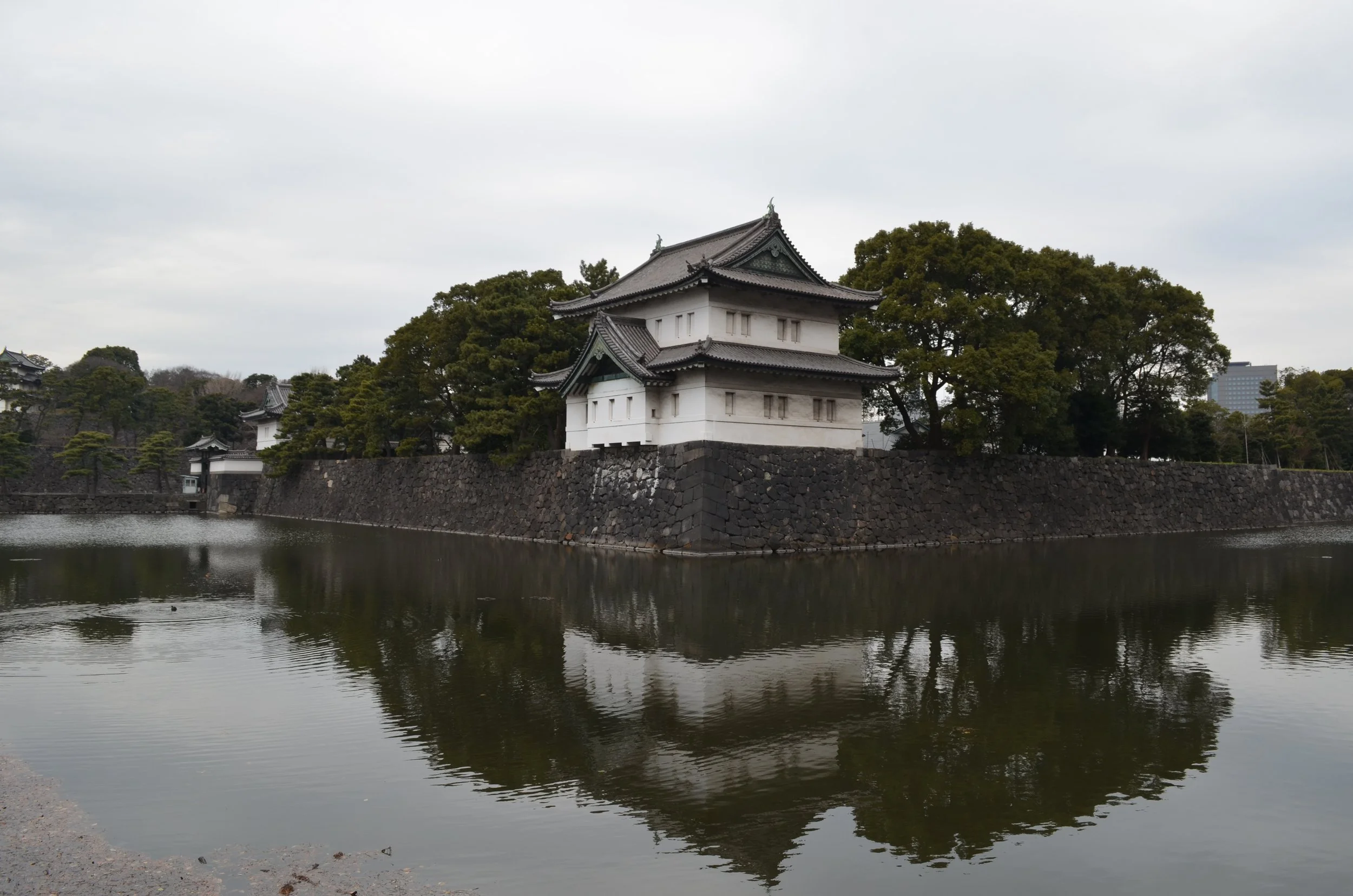 Torre tradicional japonesa junto a un muro de piedra, reflejada en un estanque, con árboles y edificio en el fondo, cielo nublado.