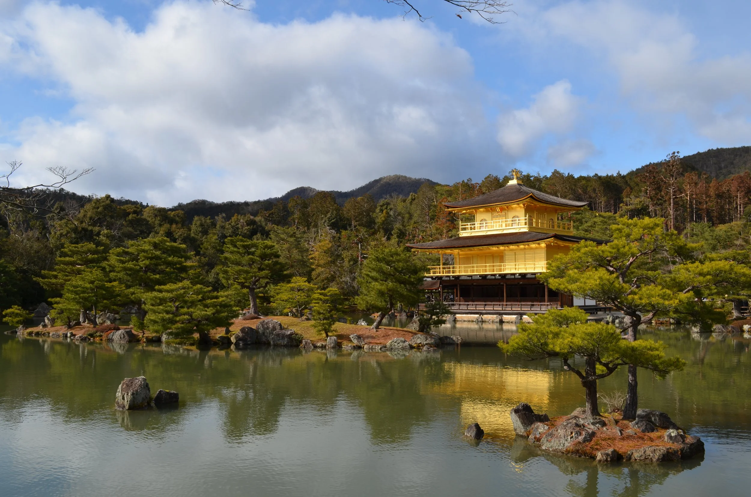 Vista del Pabellón Dorado en Kioto, Japón, rodeado de árboles verdes y un lago con rocas, con montañas y cielo nublado al fondo.