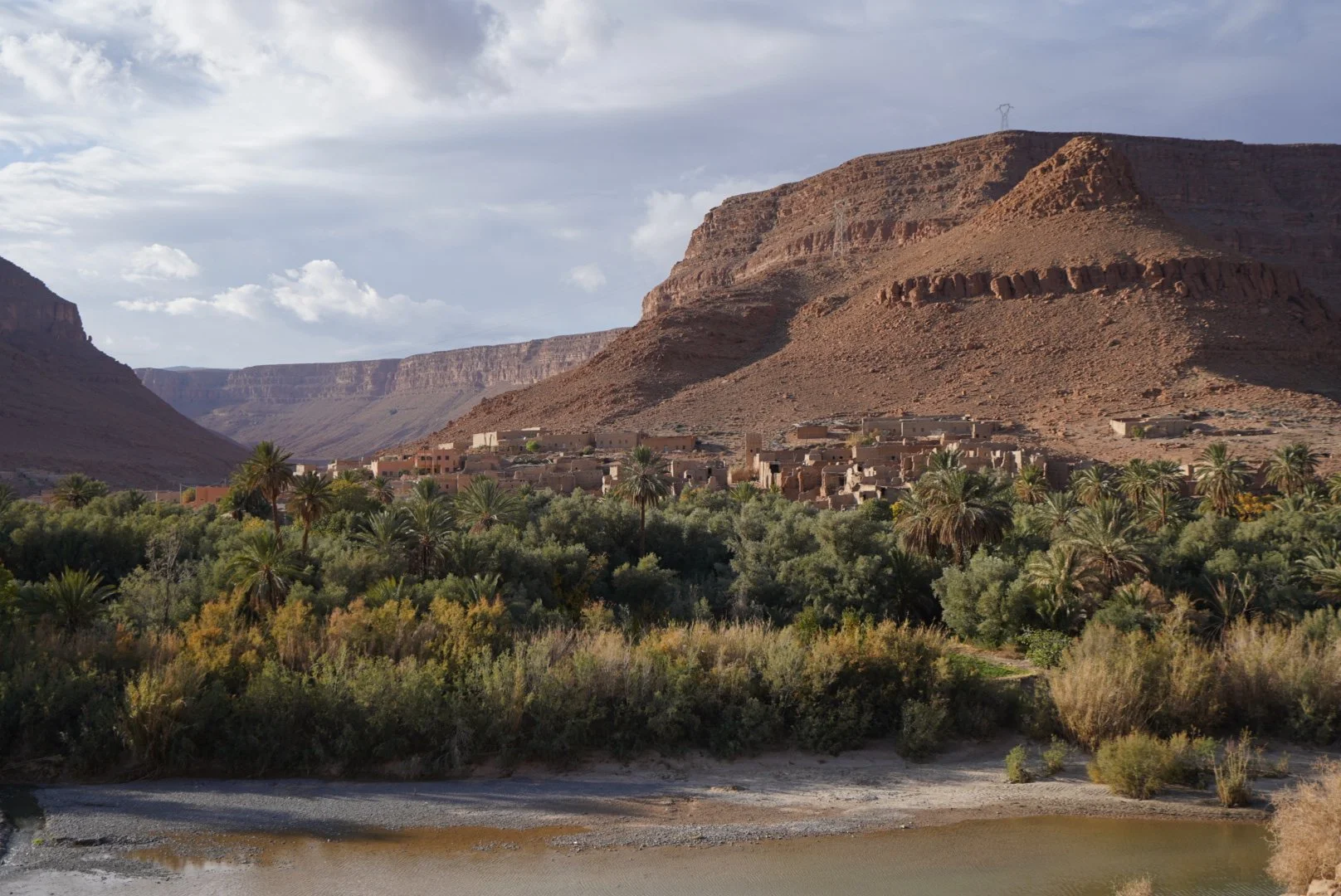 Paisaje del desierto con montañas rocosas, un oasis con palmeras y un pequeño pueblo en las faldas de las montañas.