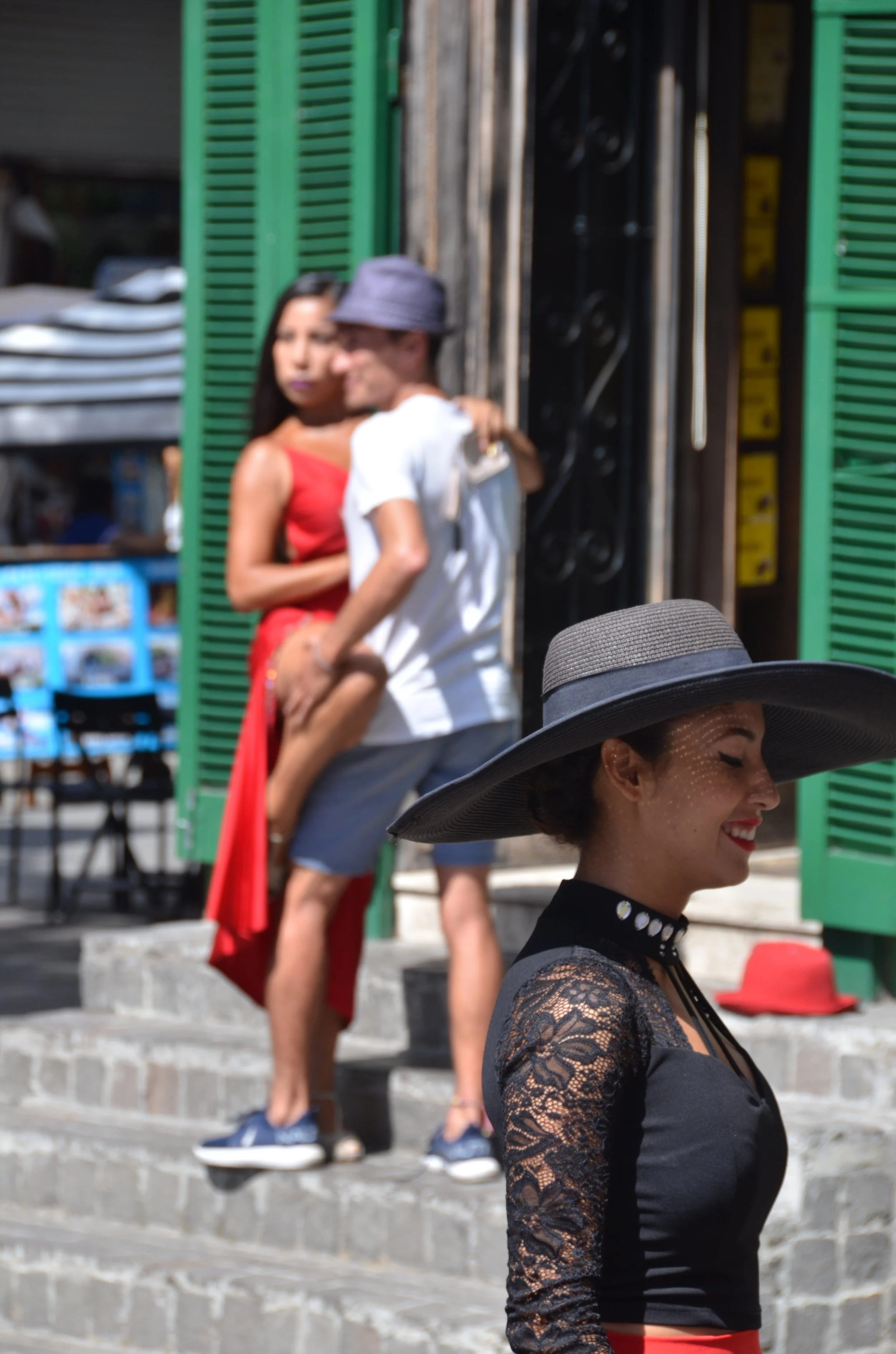 Mujer con sombrero grande sonriendo en primer plano y, en el fondo, una pareja abrazada en la acera frente a una tienda con persianas verdes.