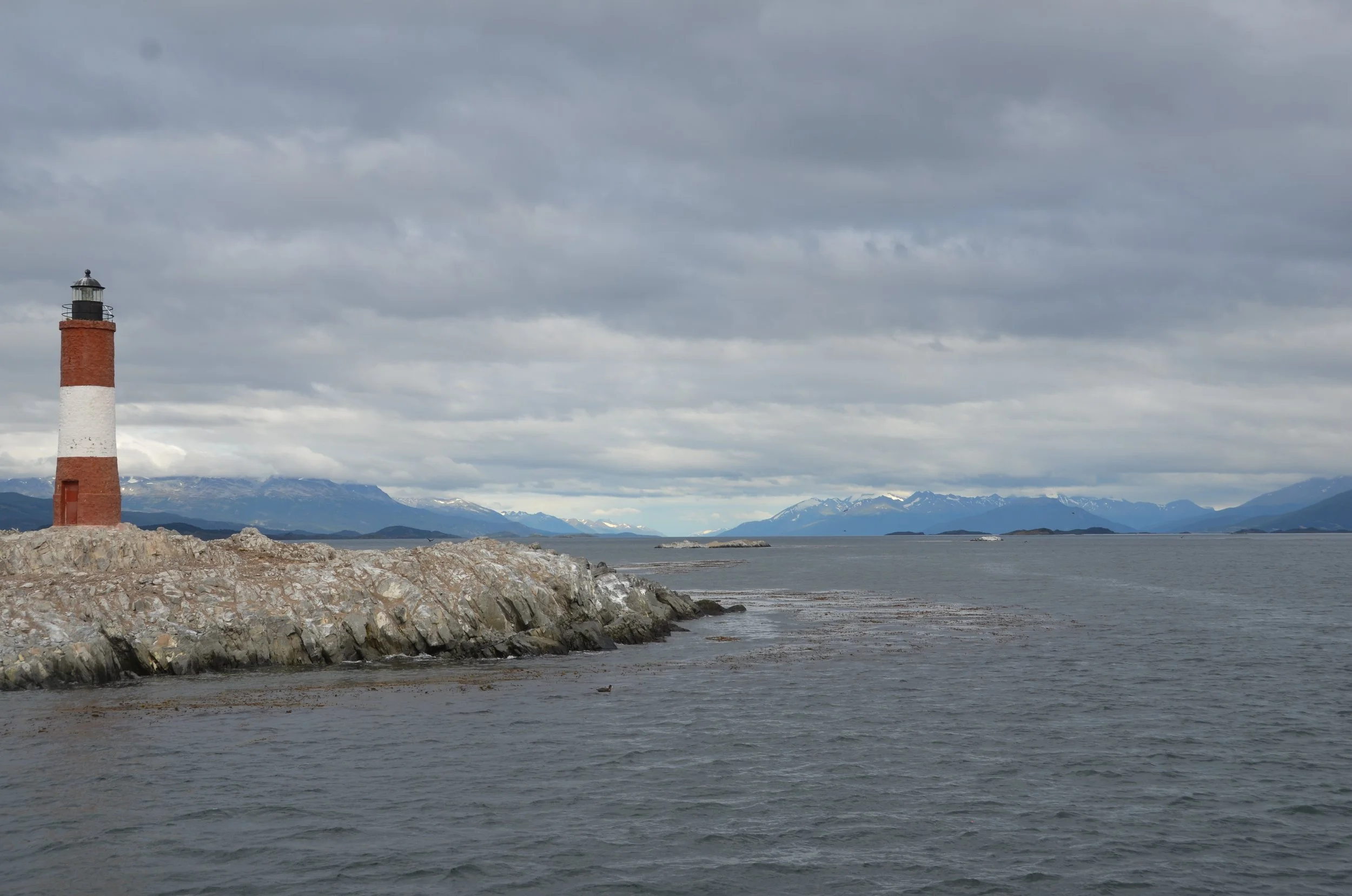 Faro de ladrillos en una roca junto al mar con montañas y un cielo nublado en el fondo.