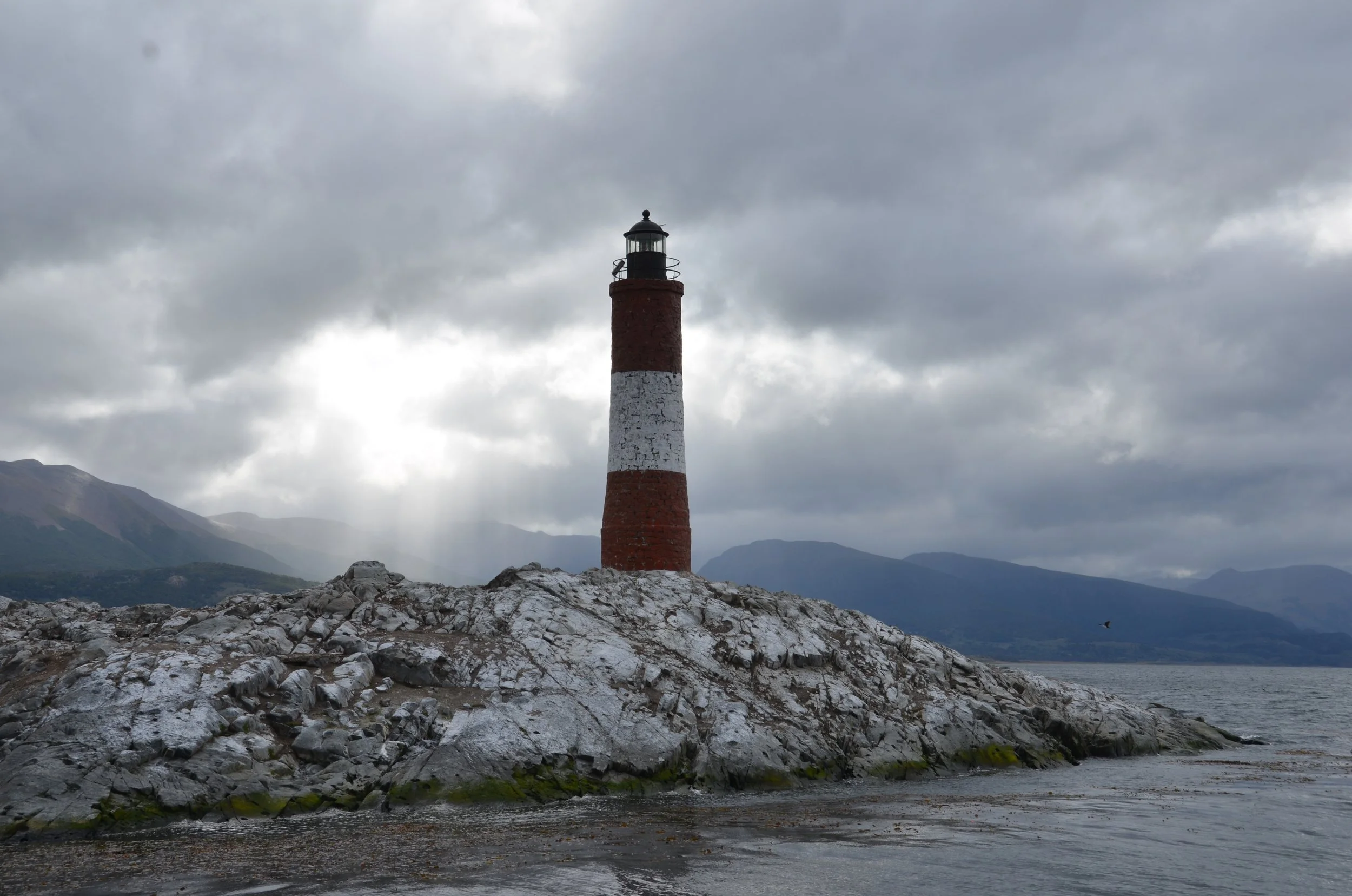 Faro de ladrillos rojos y blancos en una roca junto al mar, con montañas nubladas en el fondo, cielo nublado y bruma en el ambiente.
