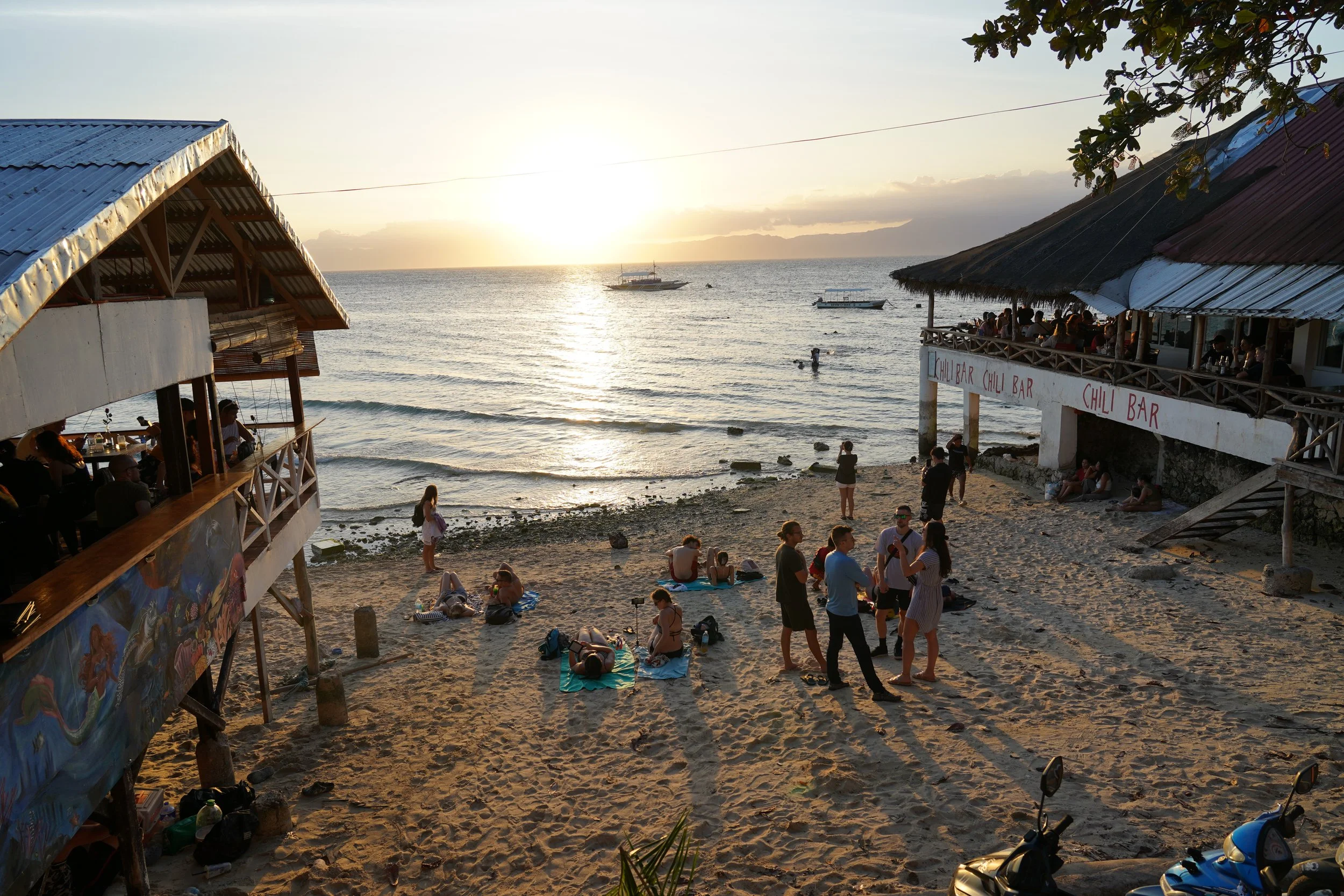 Playa al atardecer con gente, cabañas con terrazas, un bar llamado 'Chili Bar', personas bañándose en el mar, motos en la arena, y barcos en el horizonte.