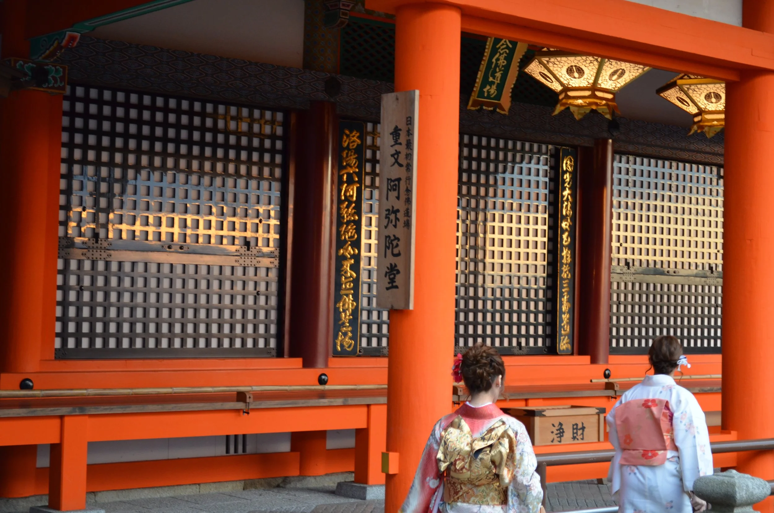 Dos mujeres con kimono tradicional en un templo japonés con puertas rojas y linternas decorativas.