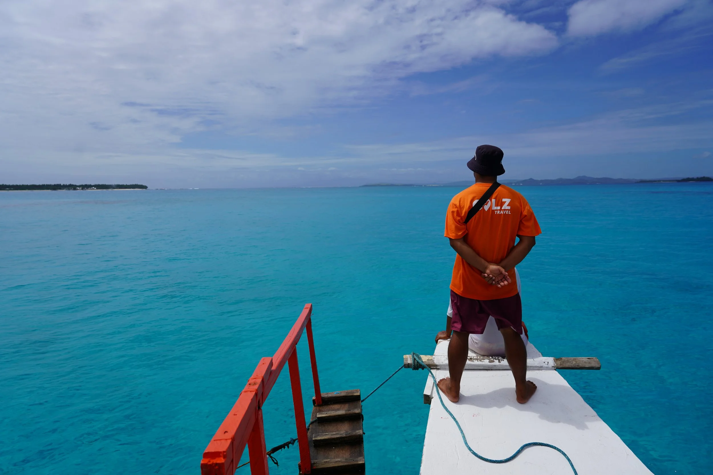 Hombre con sombrero negro y camiseta naranja de viaje en un muelle de madera, mirando hacia el mar azul con cielo parcialmente nublado.