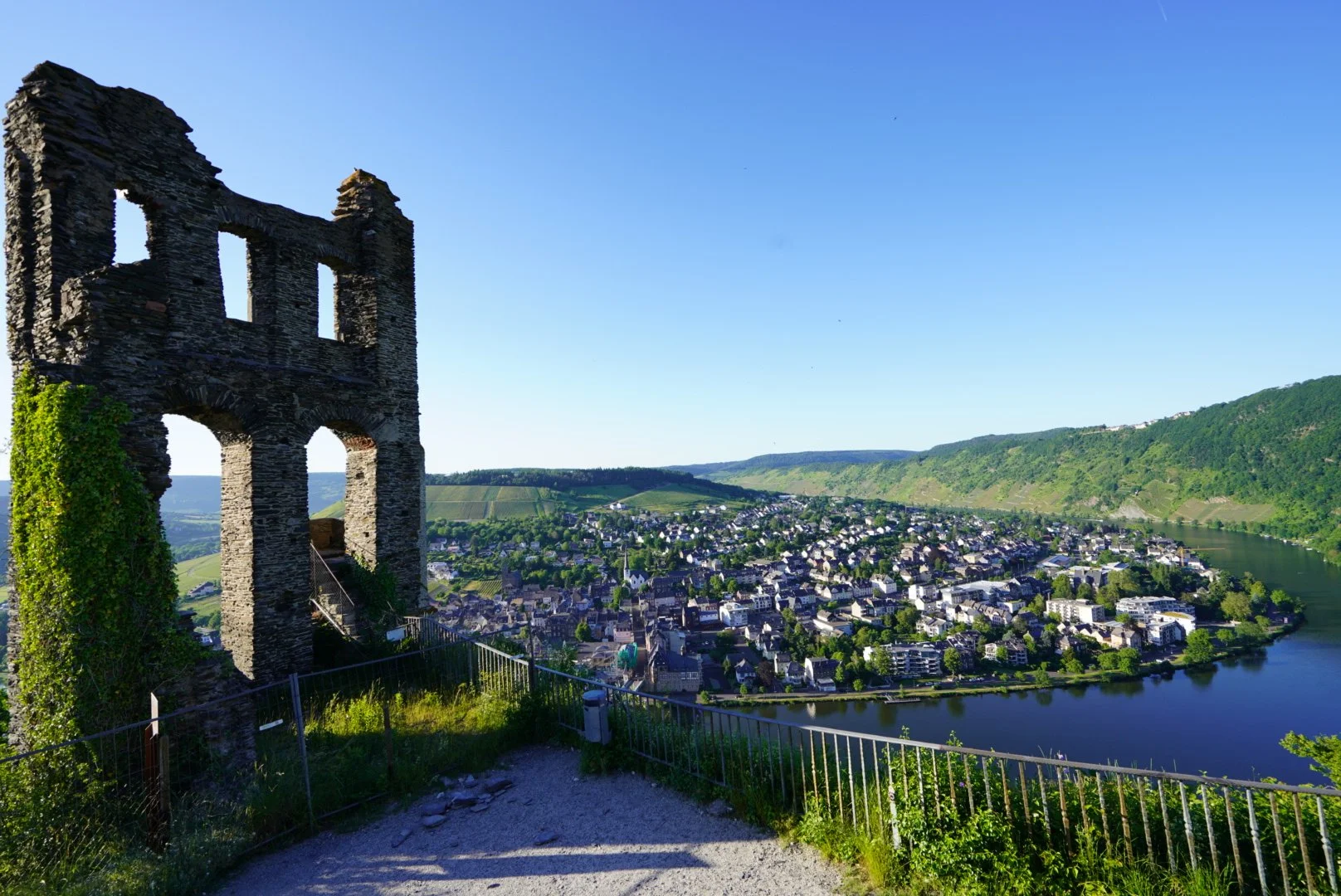 Ruinas de un edificio antiguo en un acantilado con vista al río y pueblo en el valle, rodeado de colinas verdes y cielo despejado.