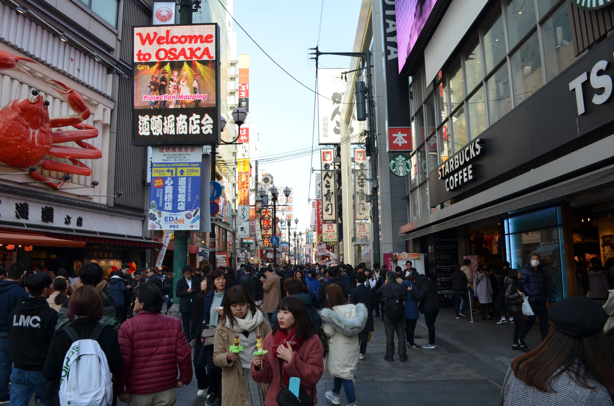 Imagen de una calle concurrida en Osaka, Japón, con muchos letreros en japonés y algunos en inglés, incluyendo un cartel que dice "Welcome to Osaka" y una tienda Starbucks con gente caminando y tiendas a ambos lados.