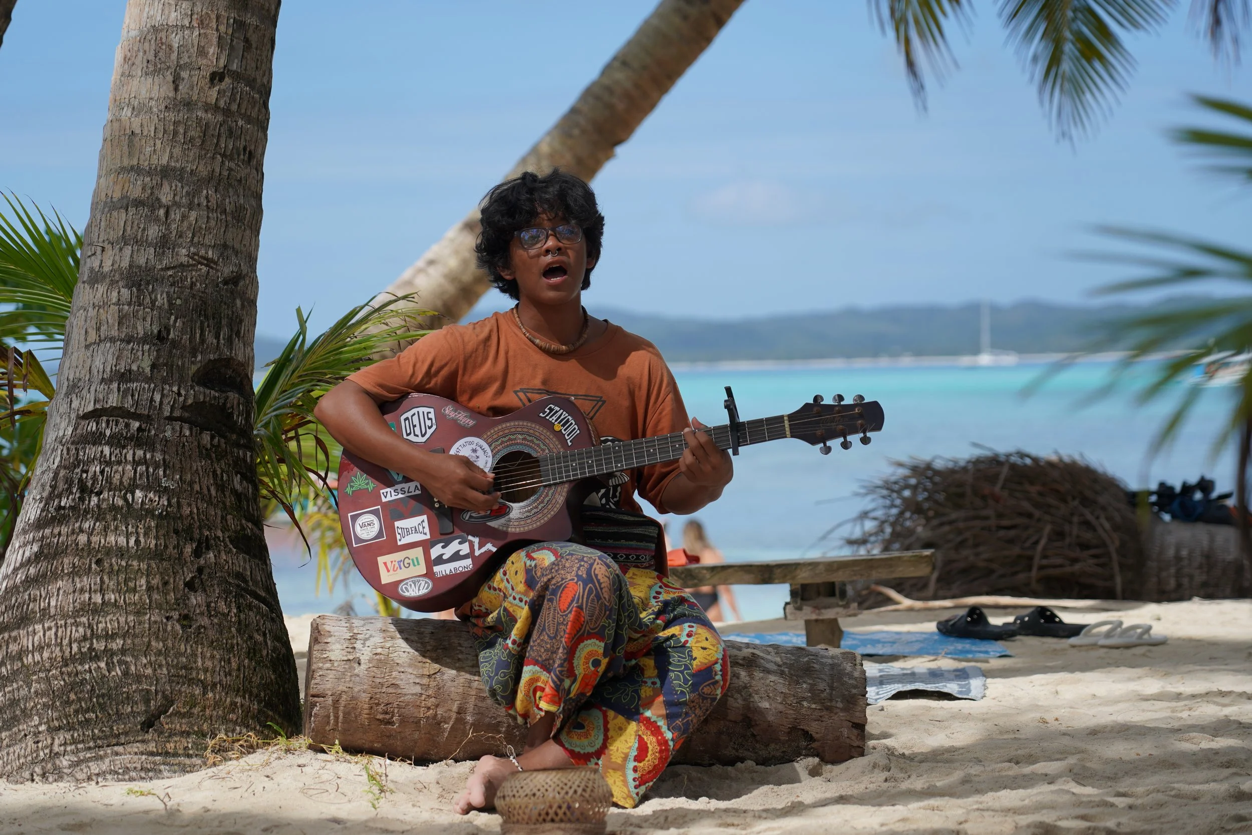 Joven tocando guitarra en la playa, sentado en tronco, rodeado de palmeras, con agua azul y cielo despejado en el fondo.