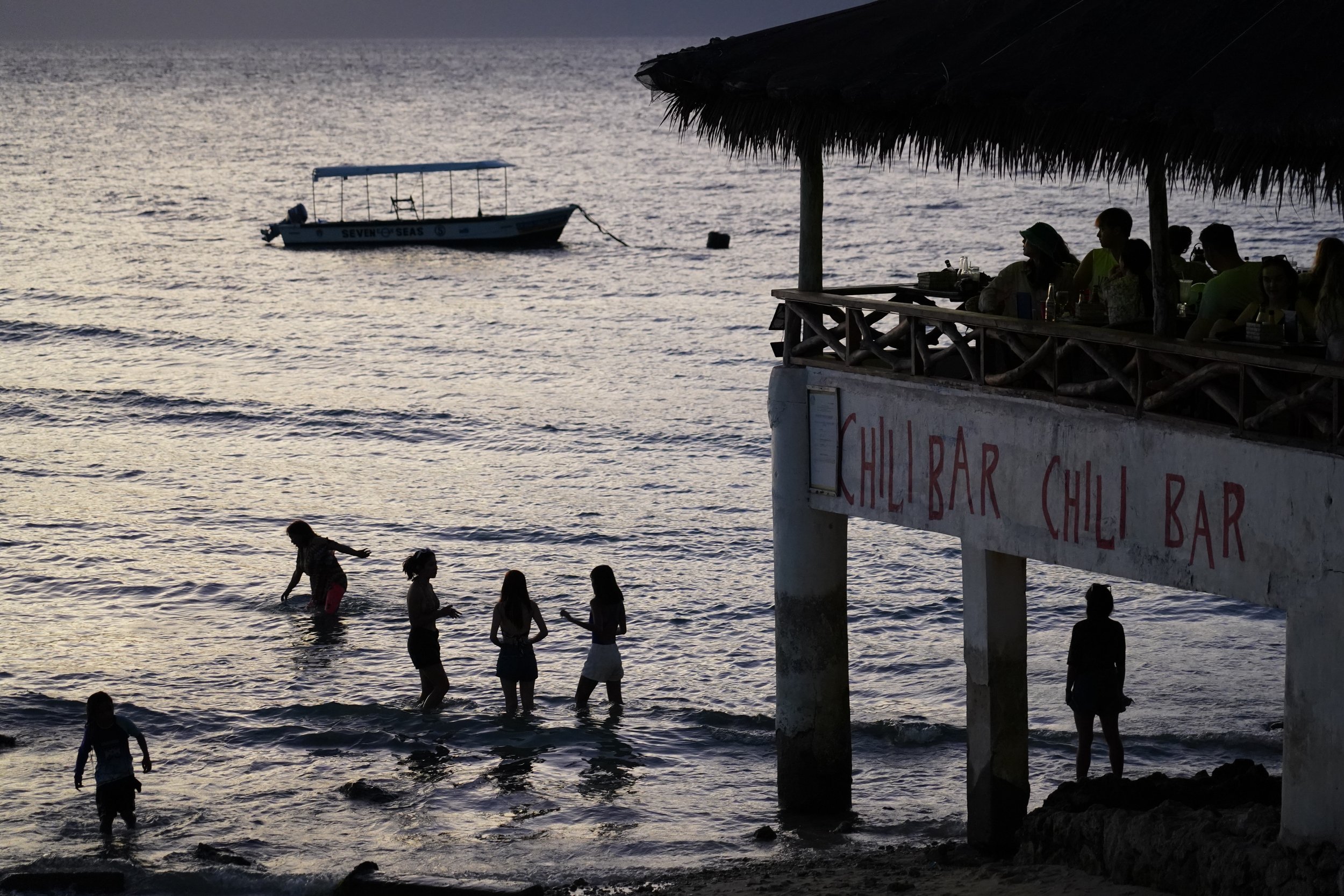 Personas en la playa y niños jugando en el agua al atardecer, mientras un bar de sushi y un bote en el mar se ven en el fondo.