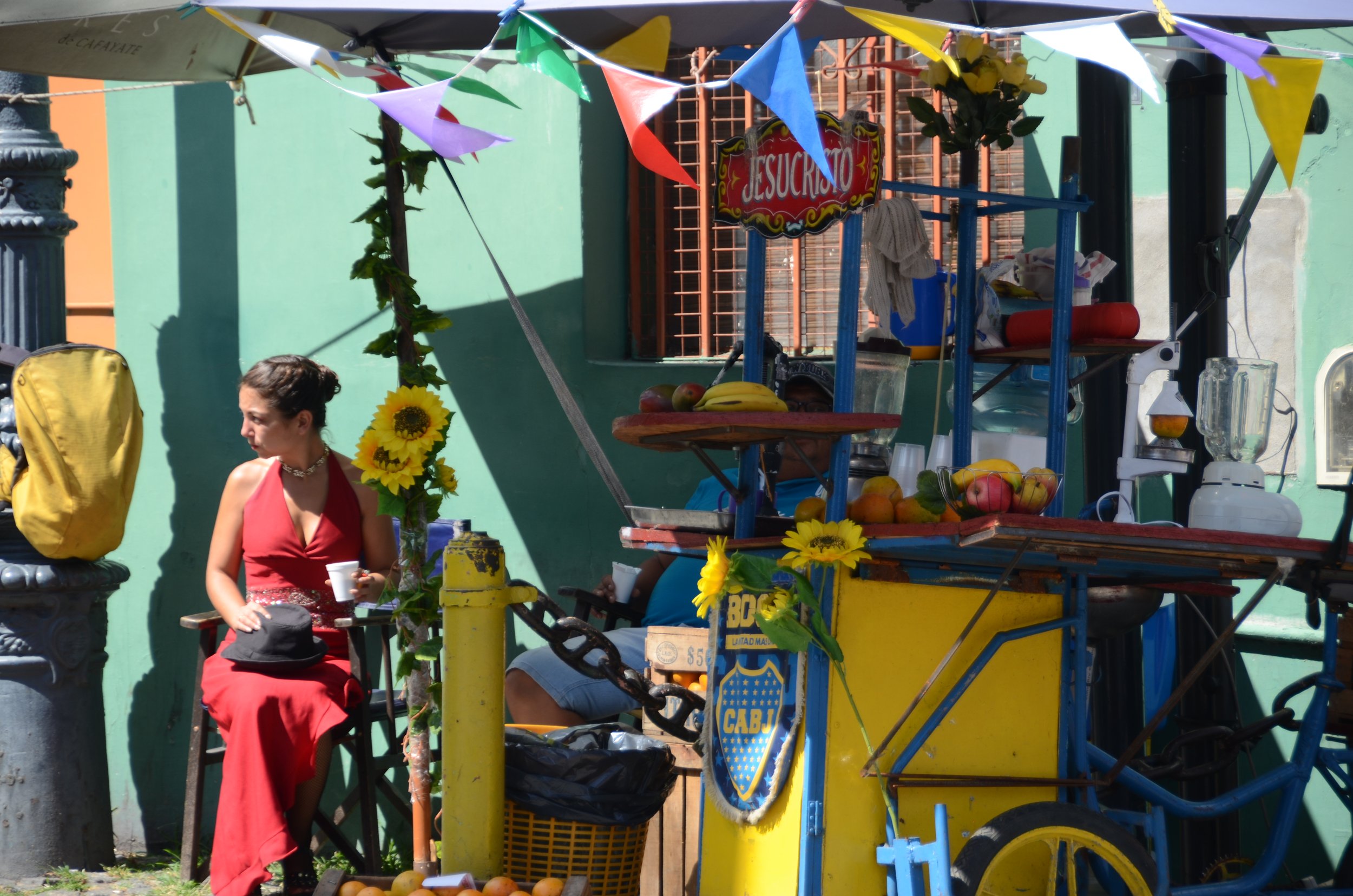 Vendedora de jugos en un puesto ambulante decorado con flores y banderines, en una calle con paredes de colores pastel, con una mujer sentada a un lado y un cartel que dice 'Jesucristo'.