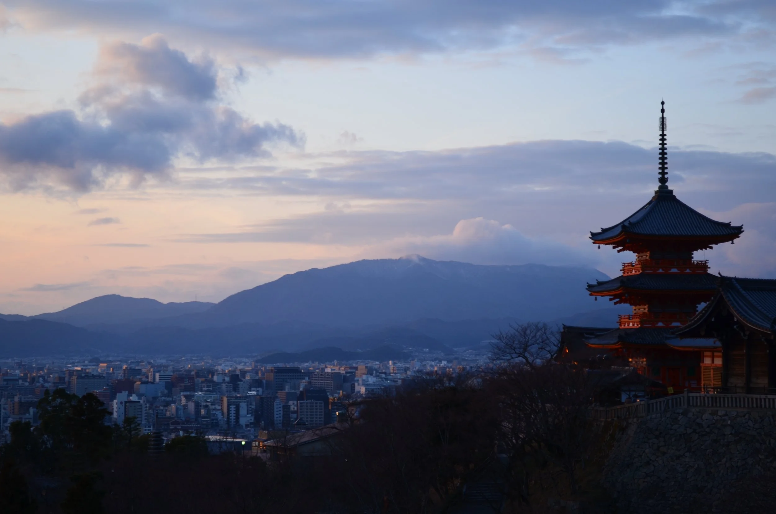 Vista panorámica de una ciudad con edificios altos y montañas al fondo, con un templo tradicional japonés en primer plano a la derecha, al atardecer.