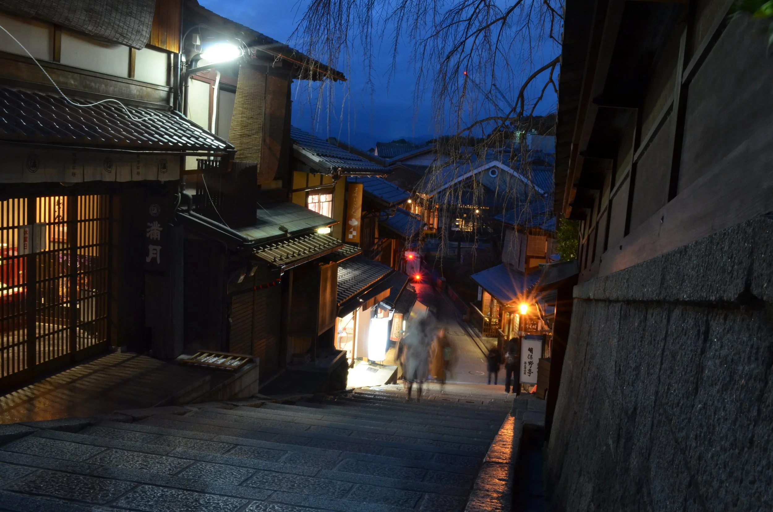 Calles empedradas en una zona histórica de noche, con casas tradicionales de madera y techos de tejas, iluminadas con faroles y luces cálidas, detalles de un ambiente tranquilo y antiguo en Japón.