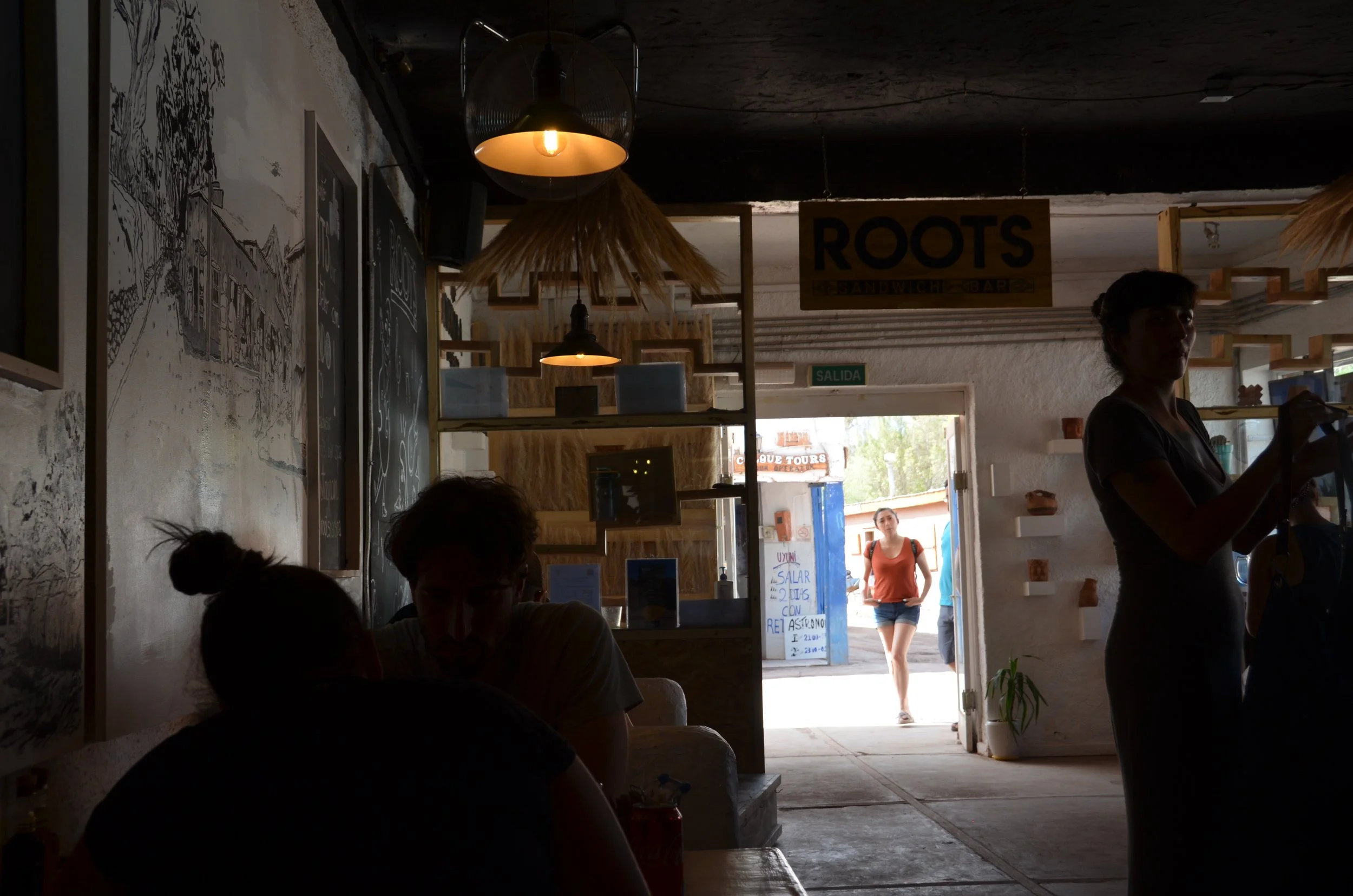 Interior de un restaurante con varias personas, algunas sentadas y otras de pie, con luz tenue y decoración moderna, con una salida al exterior donde una mujer pasa con mochila.