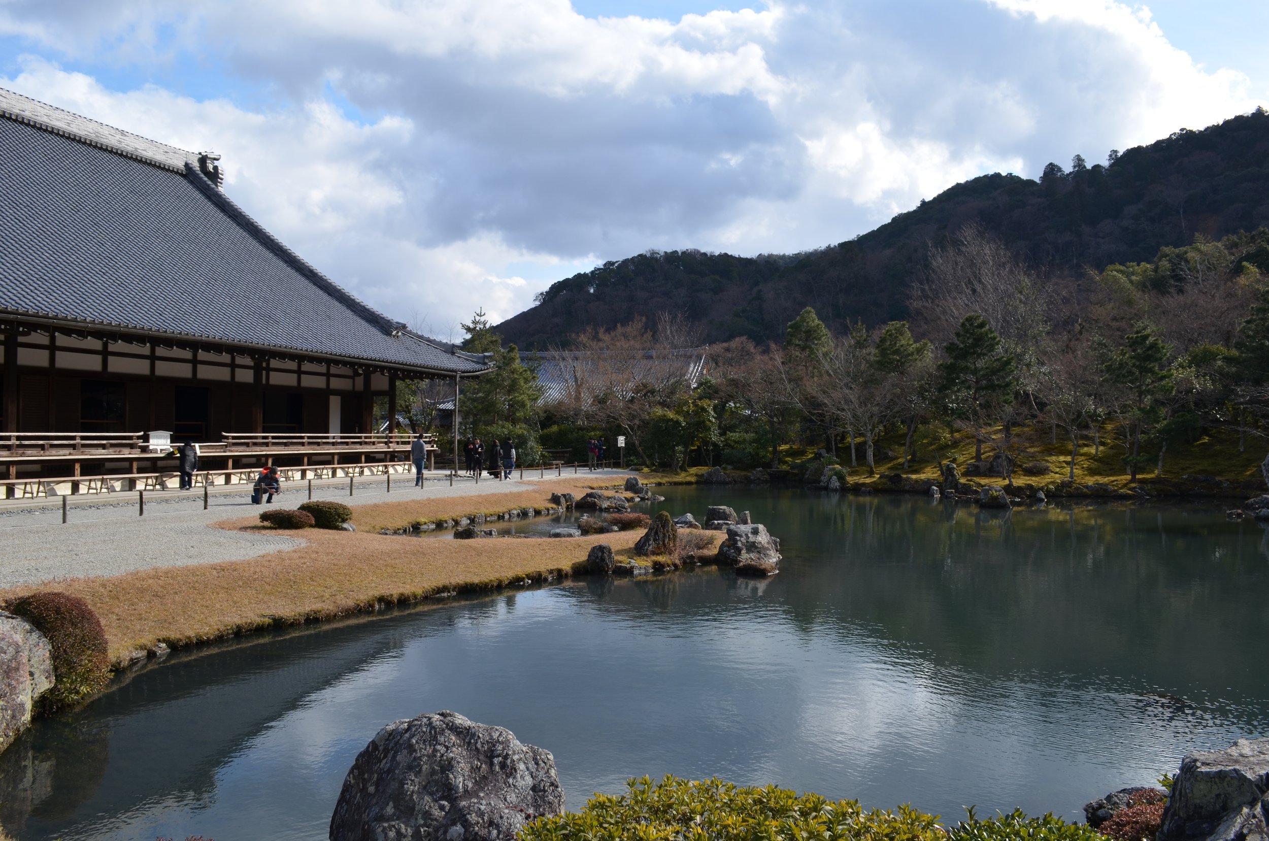 Vista de un estanque en un jardín japonés con árboles, rocas y un edificio tradicional de tejas negras, montañas al fondo y un cielo parcialmente nublado.