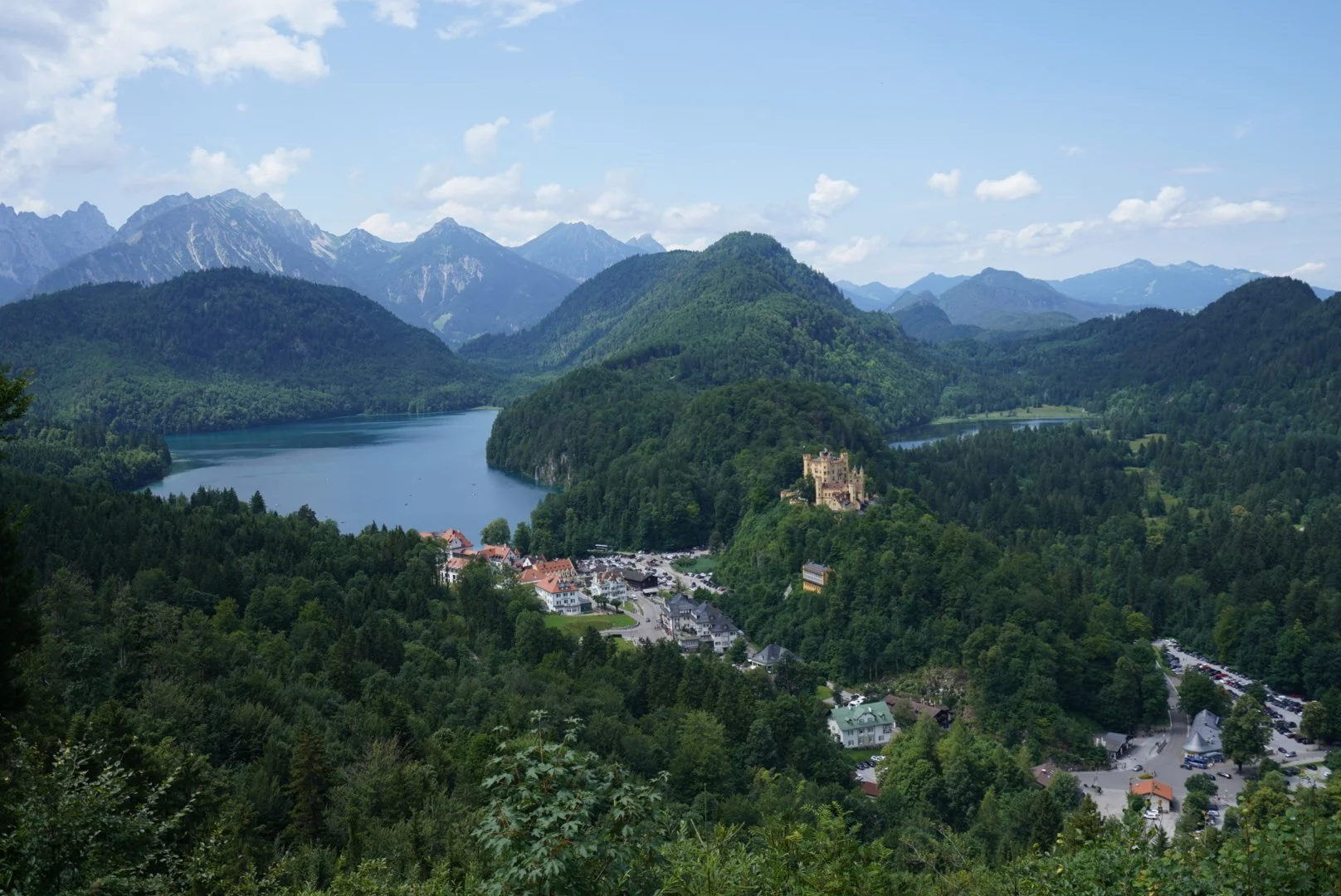Paisaje de montañas y bosques con un lago en medio, un castillo en una colina y un pueblo en la base de la colina.
