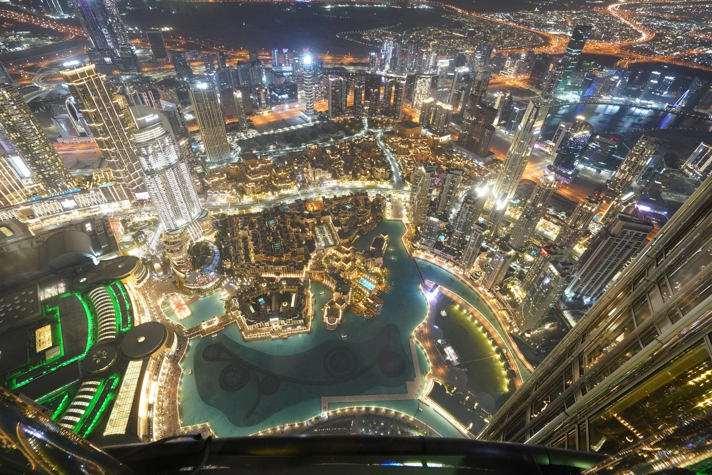 Vista aérea nocturna de Dubái desde una torre, mostrando el skyline iluminado con edificios altos y una piscina artificial en forma de姑娘 en el centro, rodeada de un río y áreas urbanas.