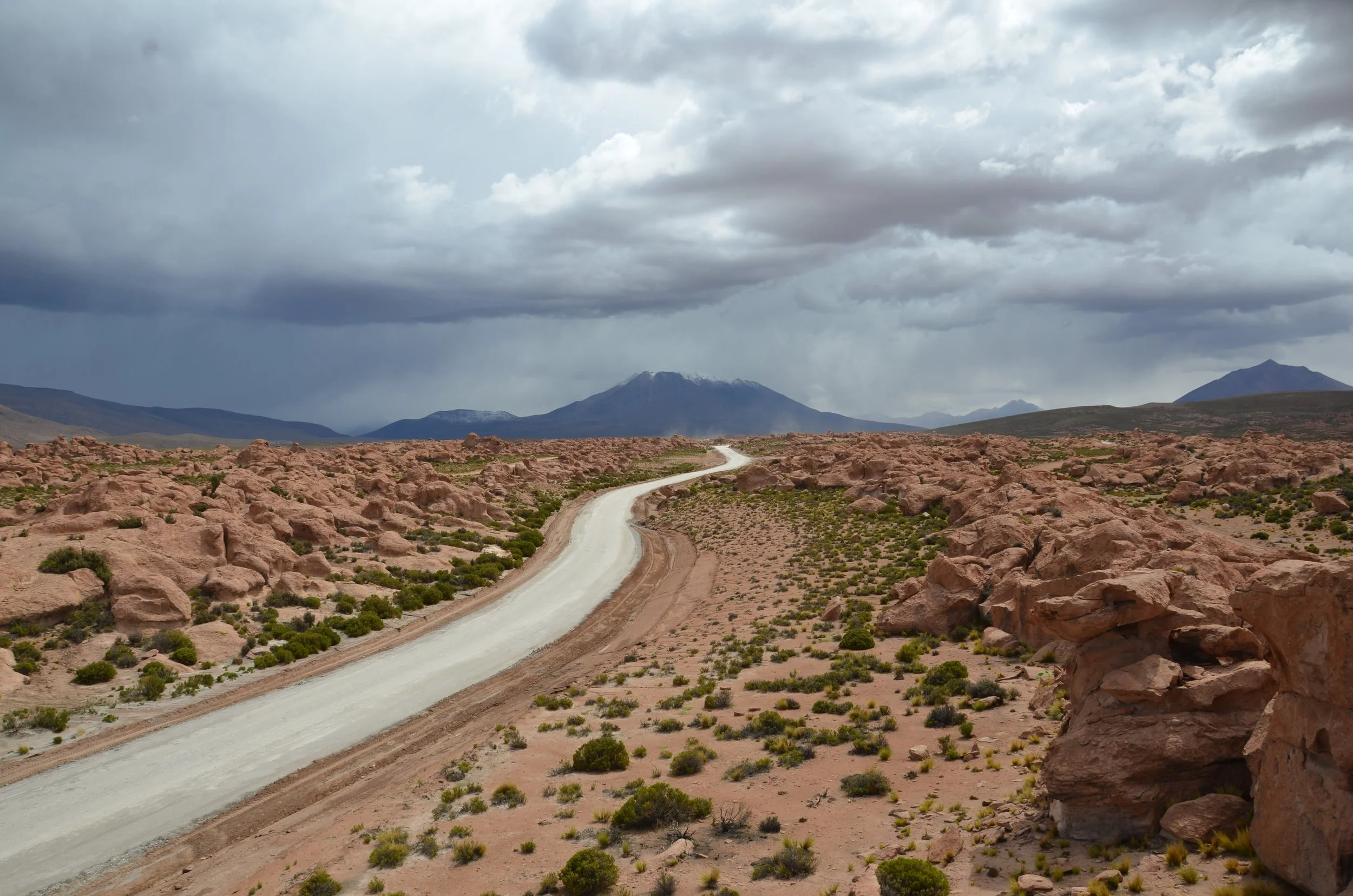 Camino de tierra en un paisaje desértico con rocas, arbustos pequeños, montañas al fondo y cielo nublado.