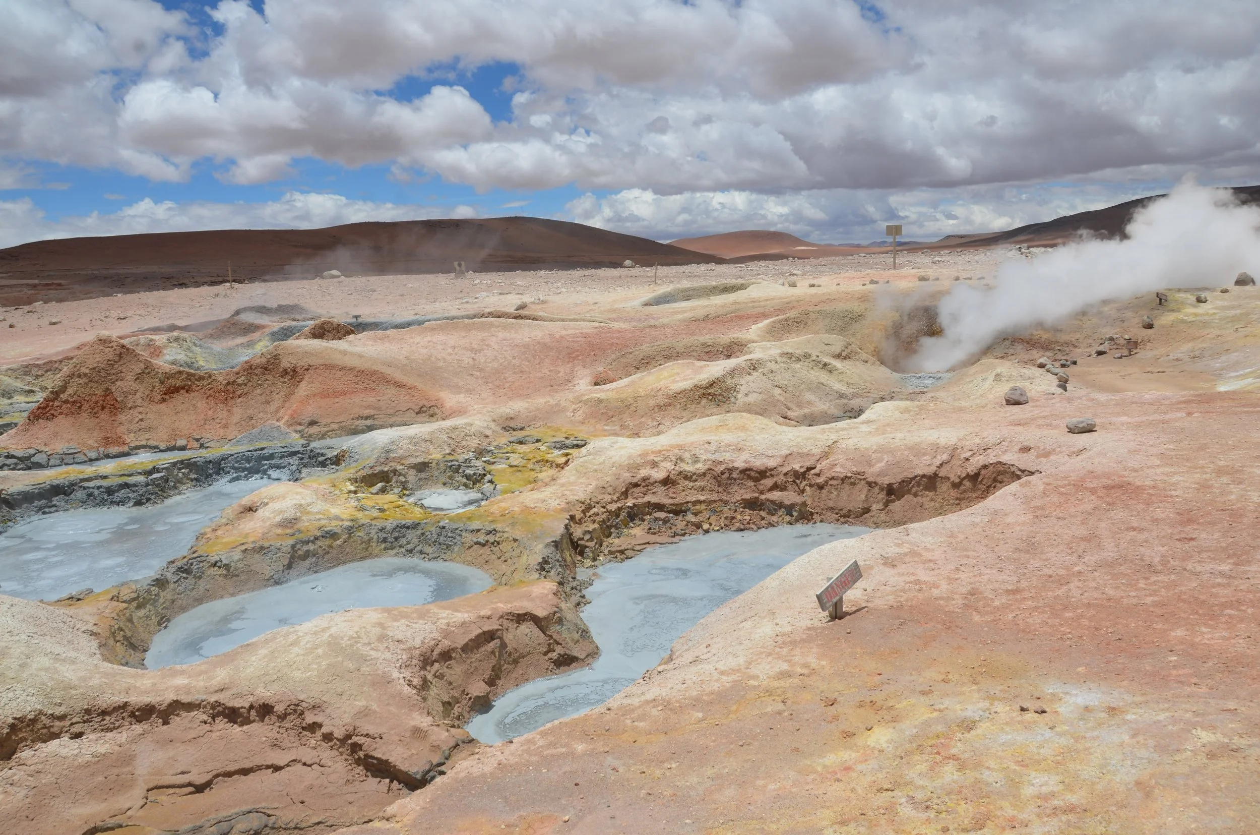 Paisaje de terreno volcánico con fumarolas y depósitos de agua caliente, en un cielo parcialmente nublado.