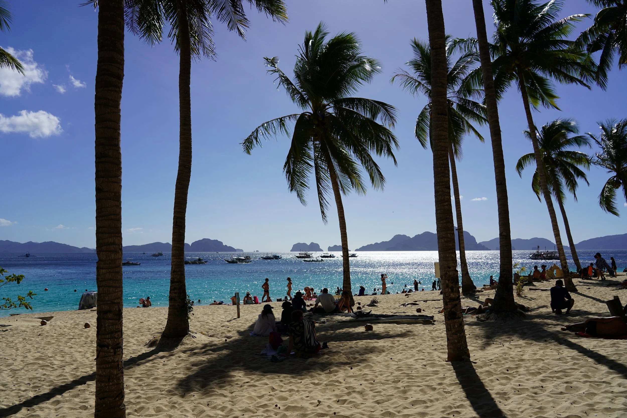 Playa con palmeras, gente en la arena y en el mar, barcos en el agua y montañas al fondo, en un día soleado y despejado.