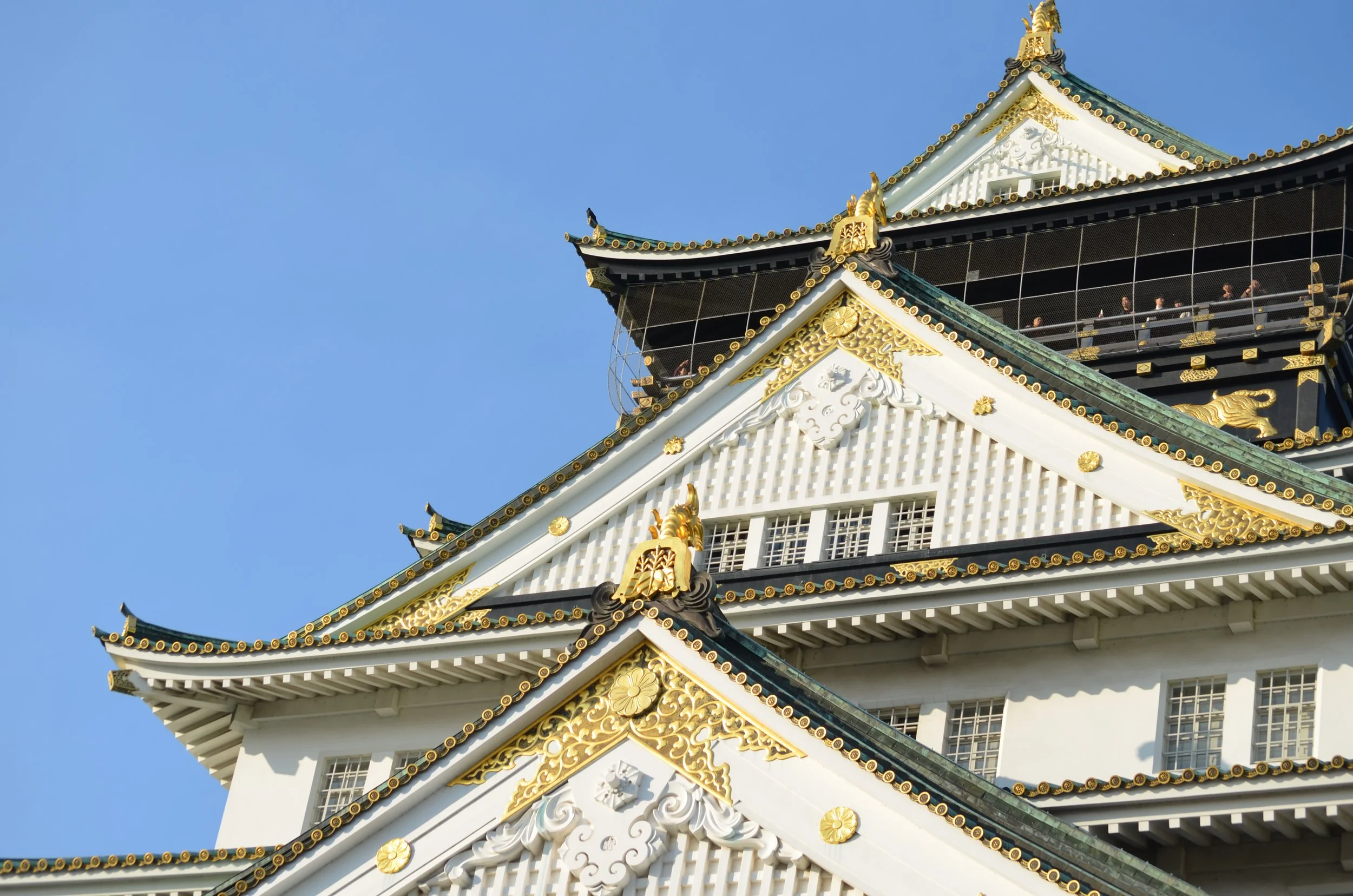 Vista del castillo de Osaka con detalles ornamentales en oro, techos inclinados y ventanas pequeñas, bajo un cielo azul claro.
