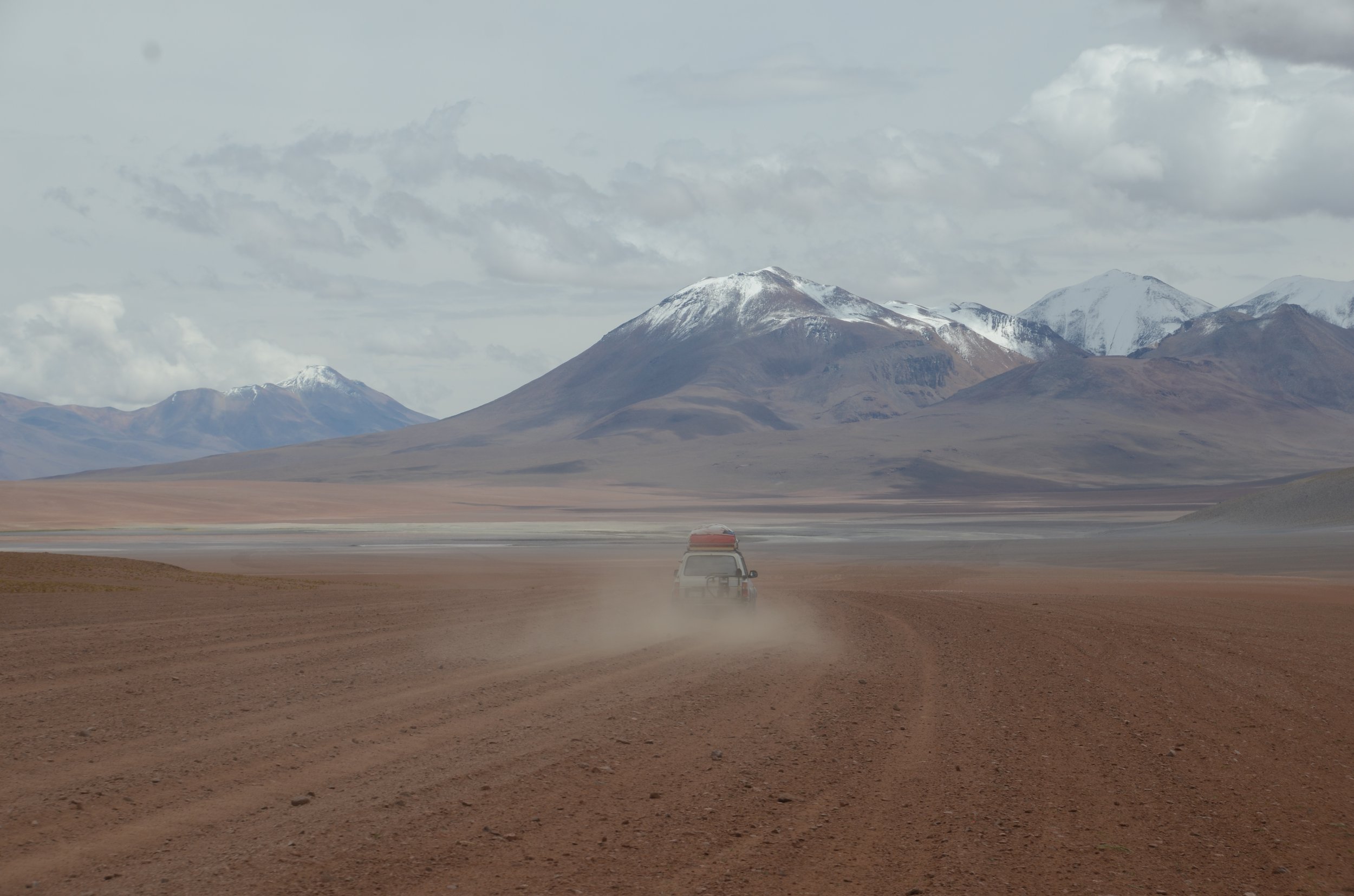 Camioneta conduciendo en un desierto rocoso con montañas nevadas y cielo nublado en el fondo.
