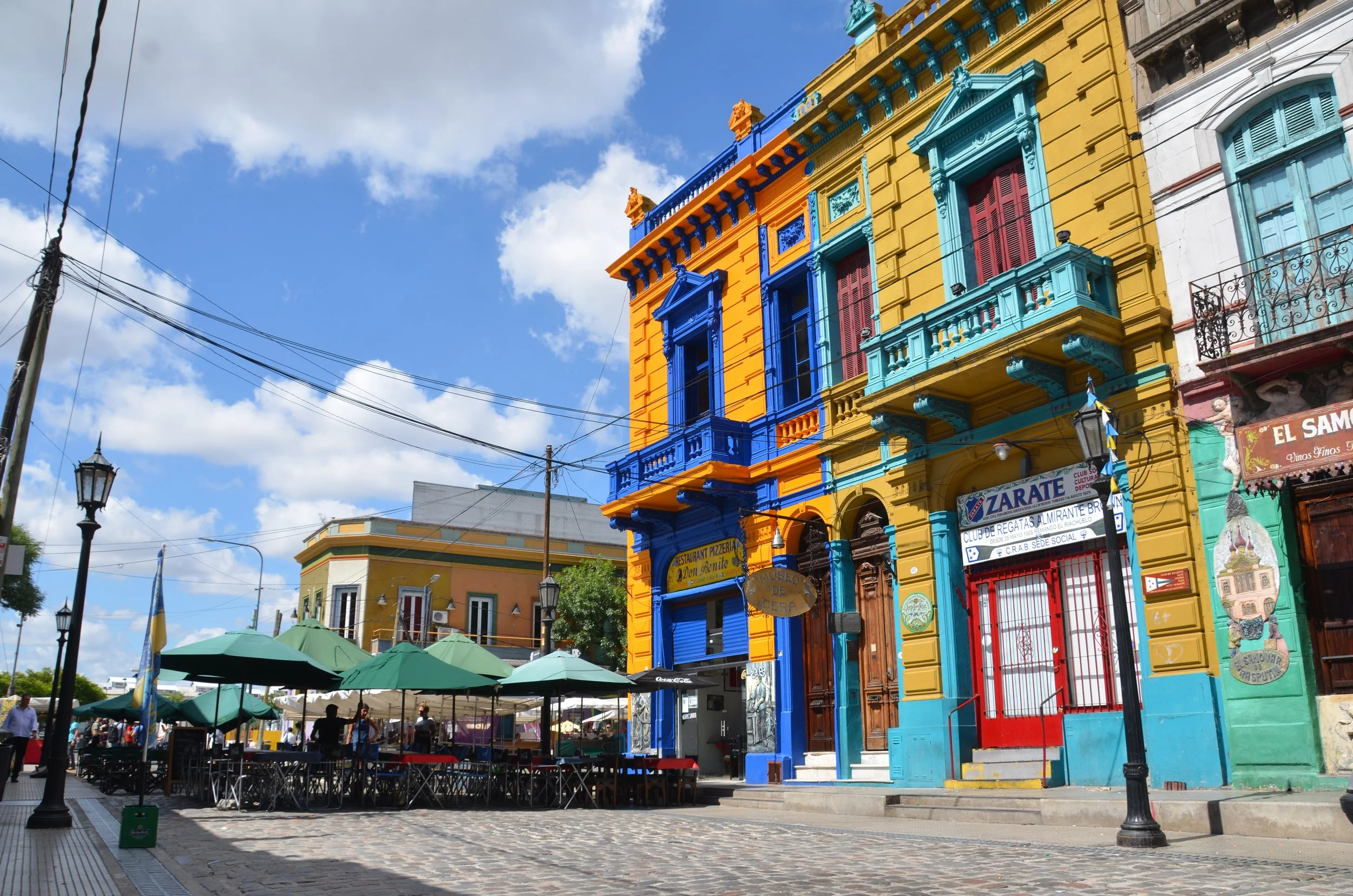 Edificios coloridos en una calle con mesas y sombrillas, cielo nublado.