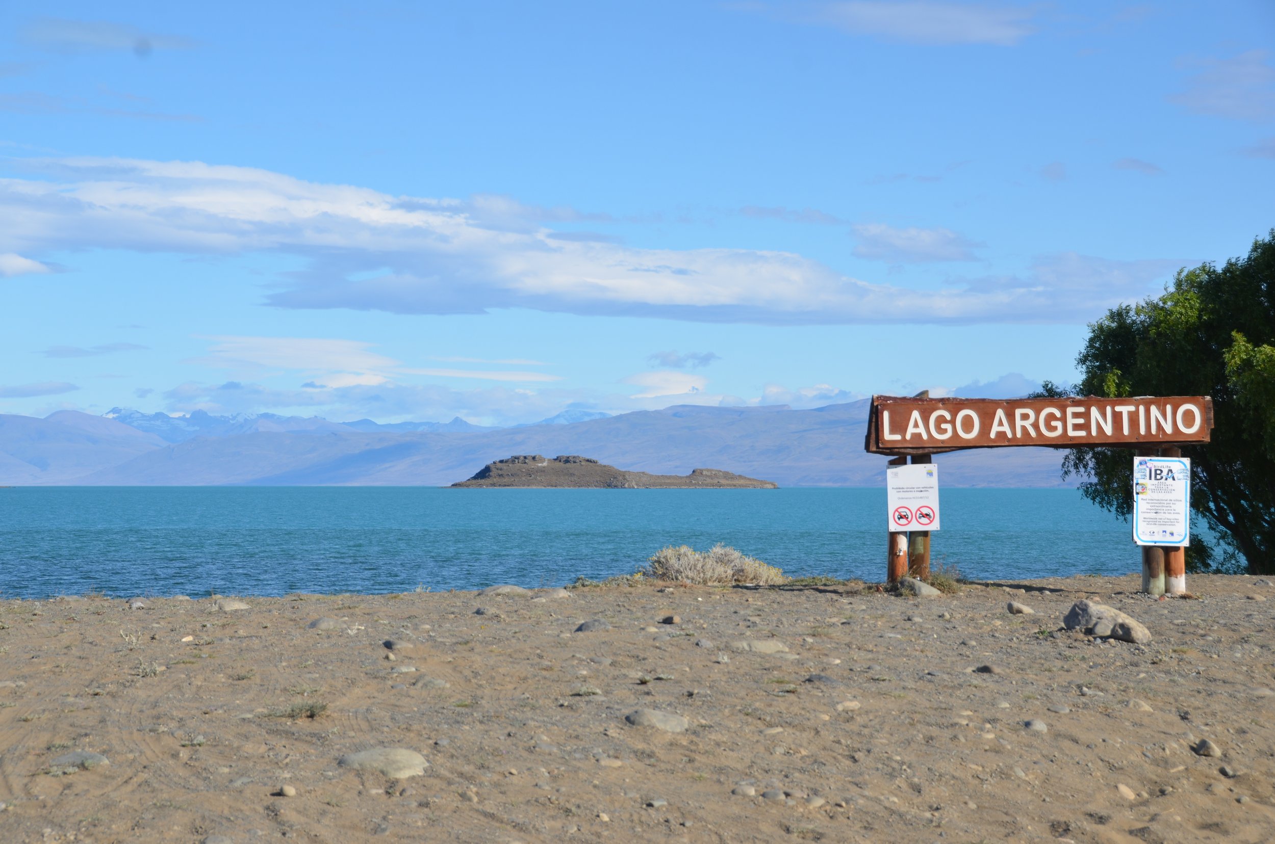 Paisaje con lago y montañas al fondo, señal que dice 'Lago Argentino' y advertencias de no nadar ni hacer fuego.