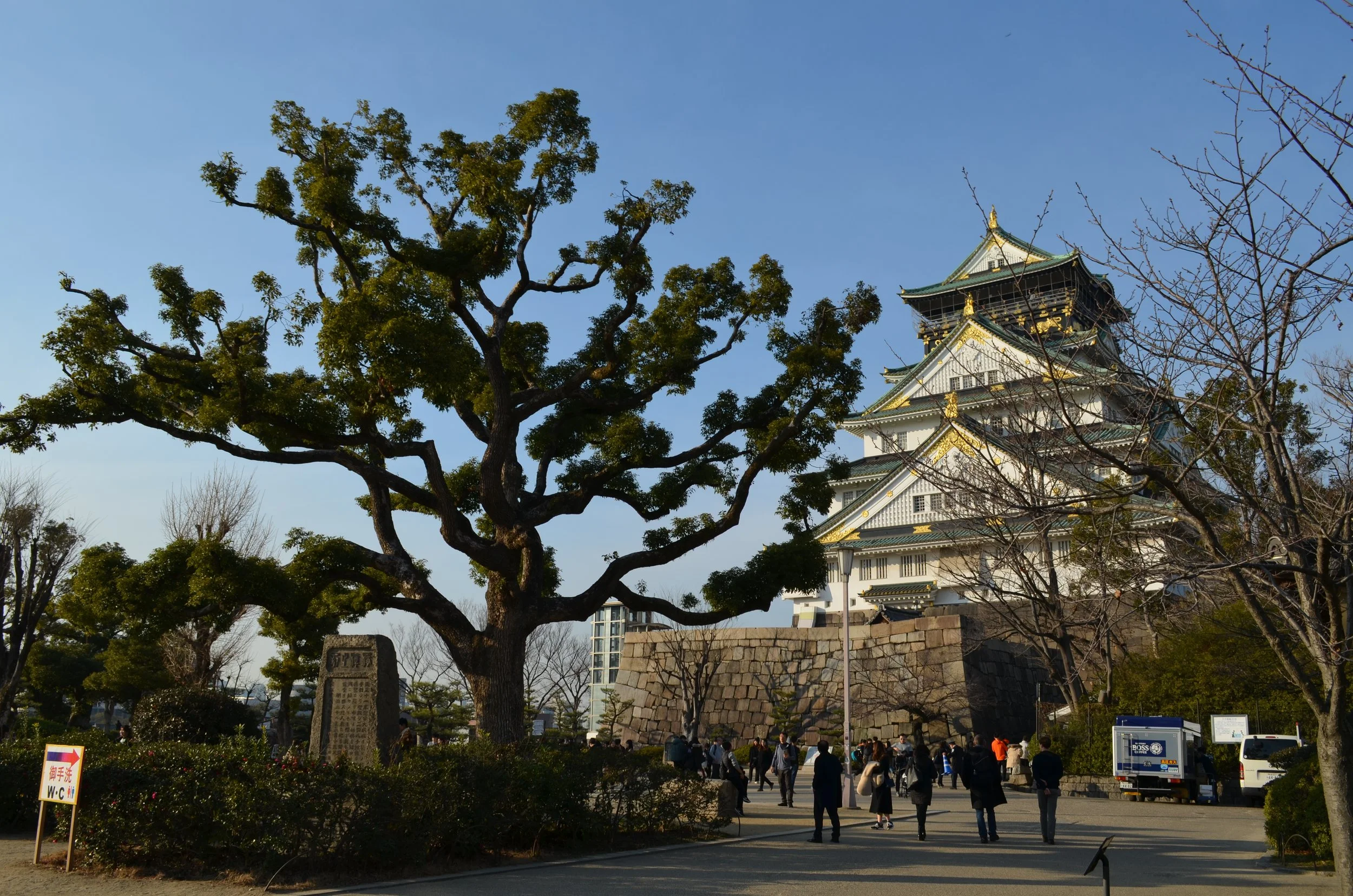 Castillo de Osaka con árboles y personas caminando en el parque cercano bajo un cielo despejado.