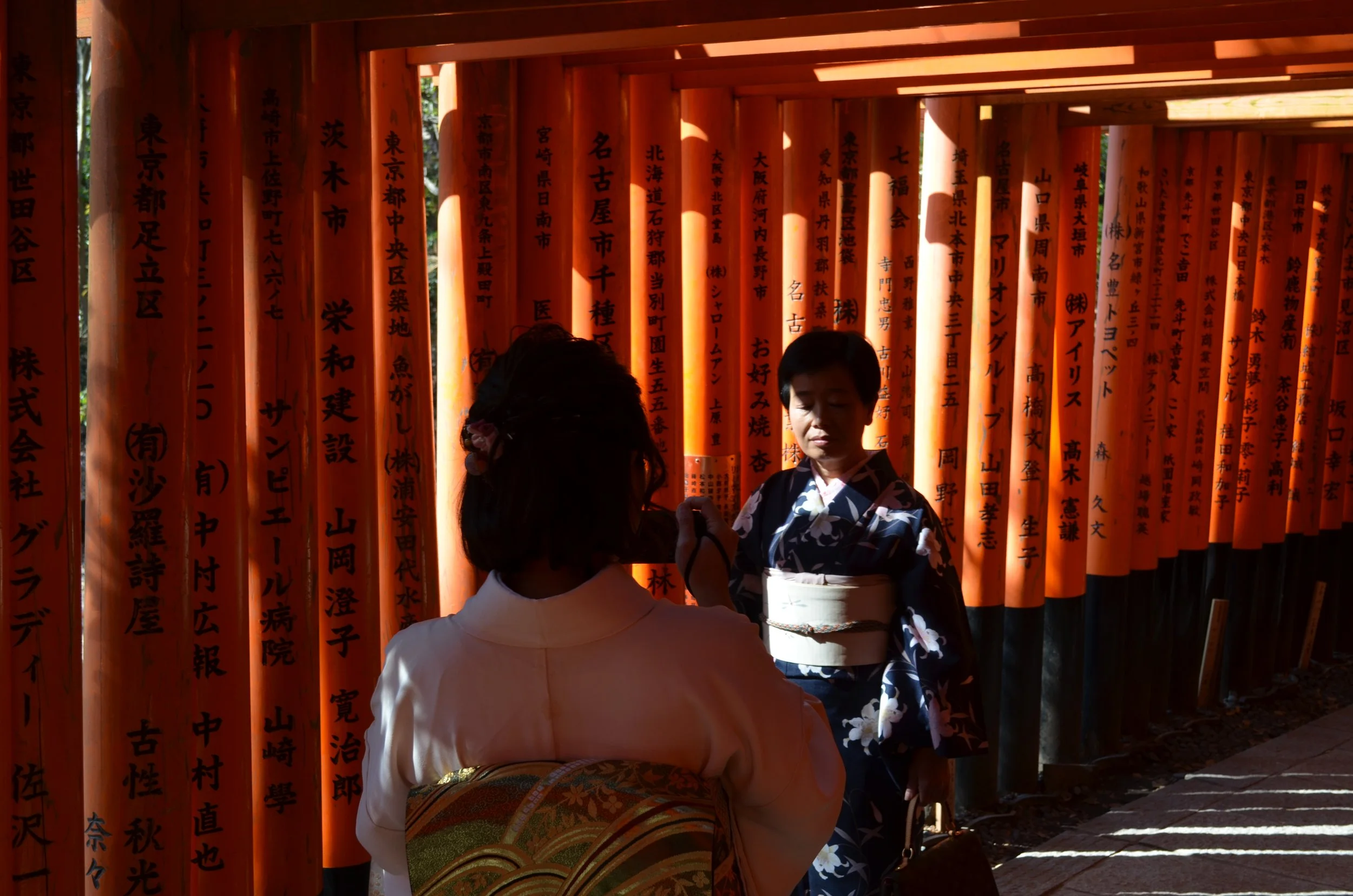 Dos mujeres con kimono interactuando frente a una fila de puertas torii de color rojo con inscripciones en japonés, en un entorno tradicional japonés.