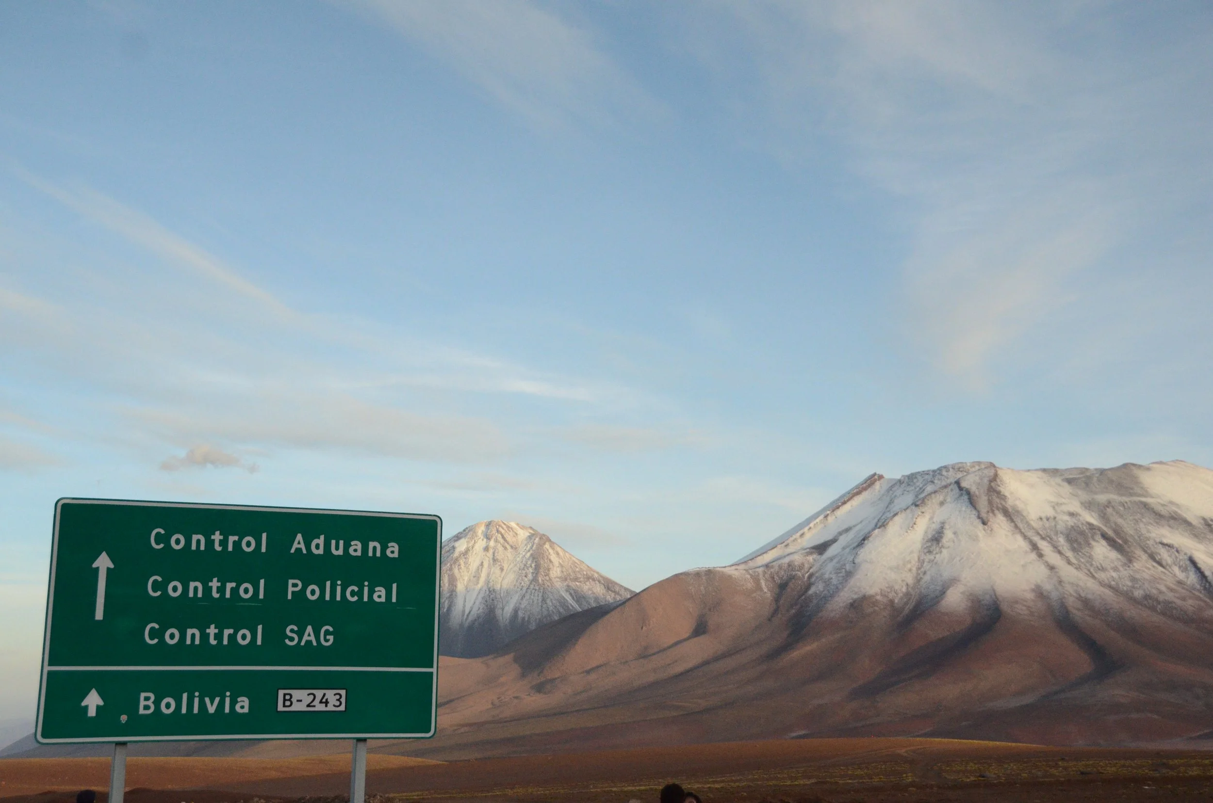 Mapa de carreteras en Bolivia con montañas nevadas en el fondo y un cartel de señalización que indica control aduanero, control policial, control SAG y dirección a Bolivia.