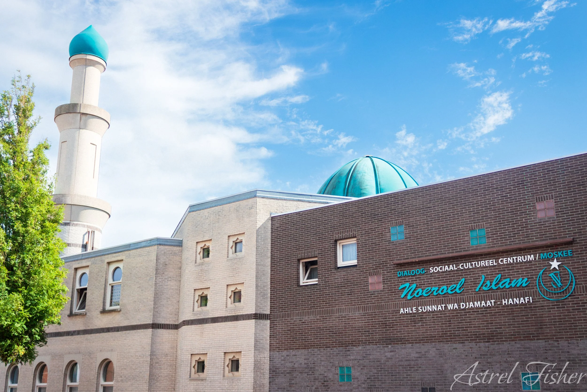View of a mosque with a tall minaret and a blue dome, situated on a city street with a brick building and a sign that reads 'Nooroe Islam.'