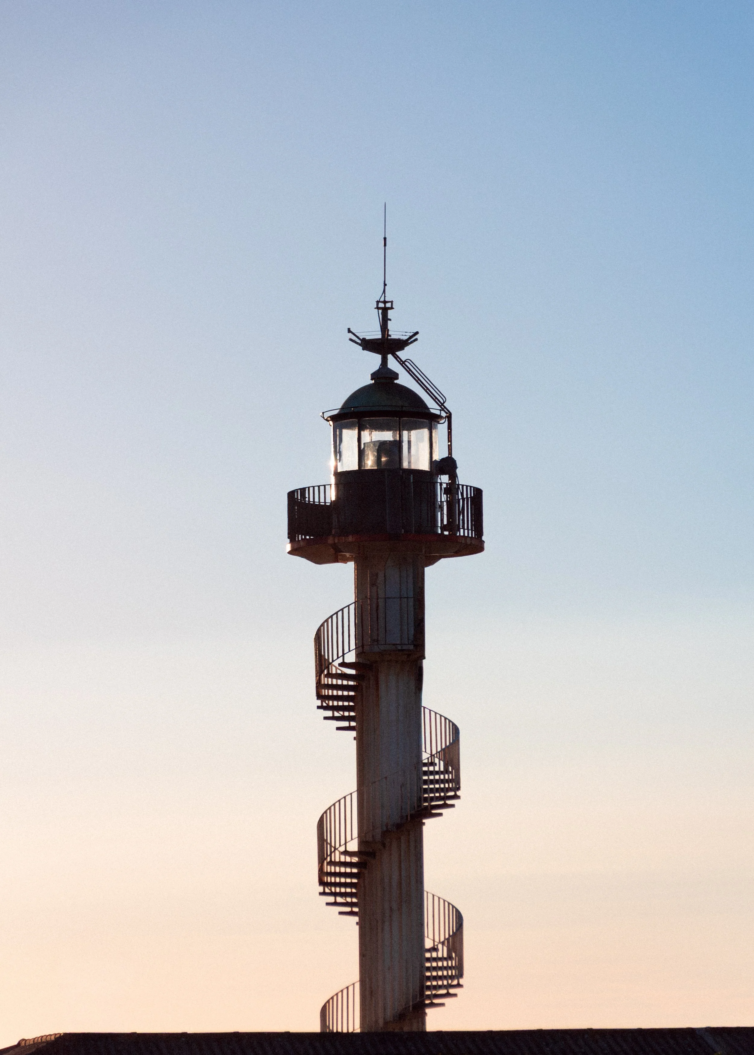 A lighthouse with a spiral staircase wrapping around its exterior, topped with a lantern room and antennas, against a clear blue sky.
