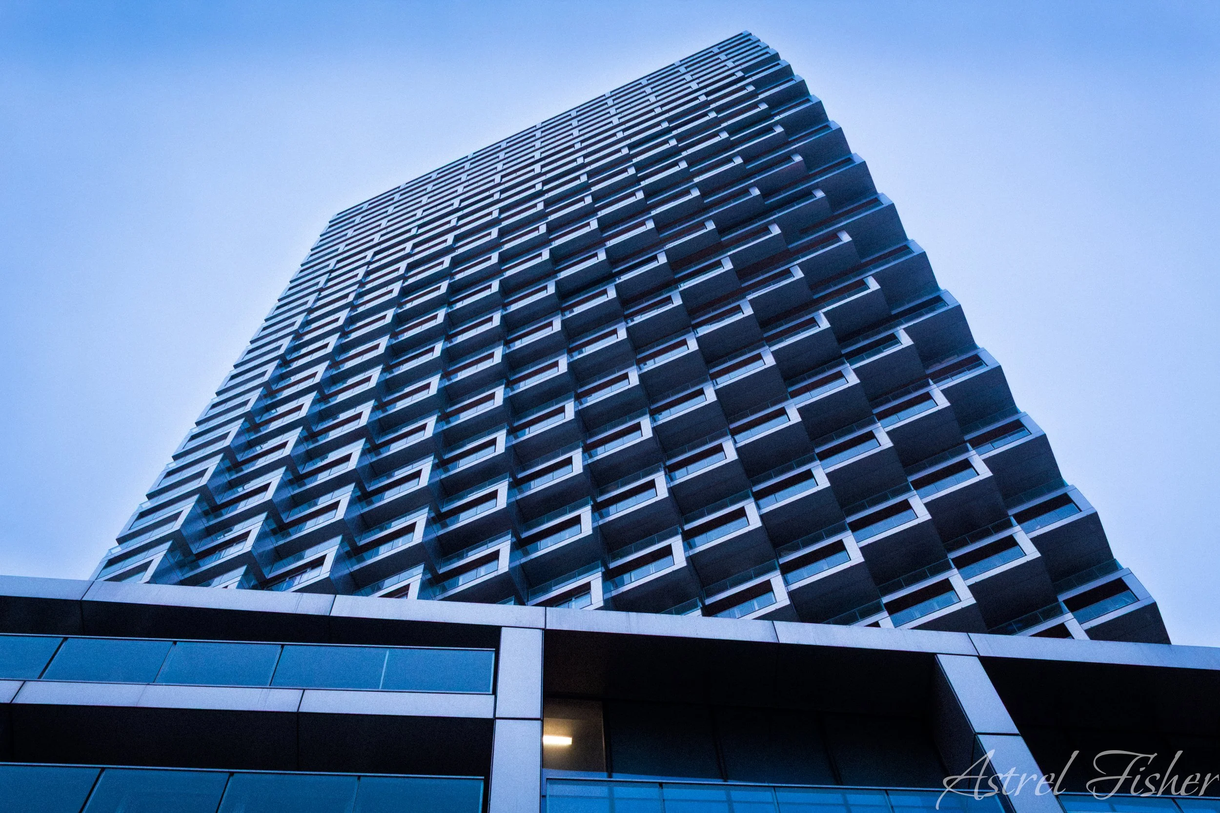 A modern high-rise building with a geometric design, featuring repeating rectangular patterns and glass windows, set against a clear blue sky.