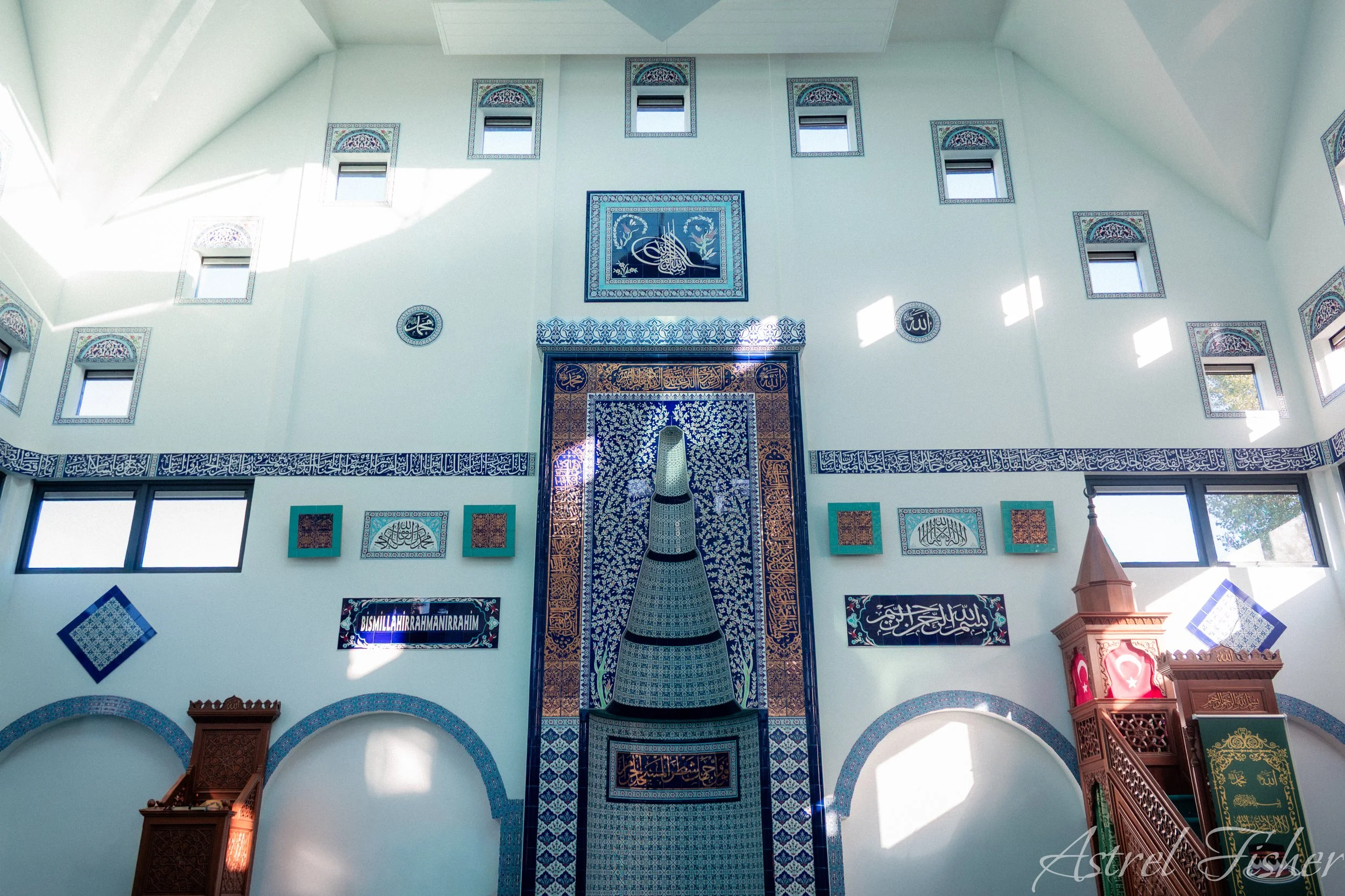 Interior of a mosque with Islamic calligraphy and decorative tiles, featuring a central stained glass window and several smaller windows, with a prayer niche and various Islamic plaques on the walls.