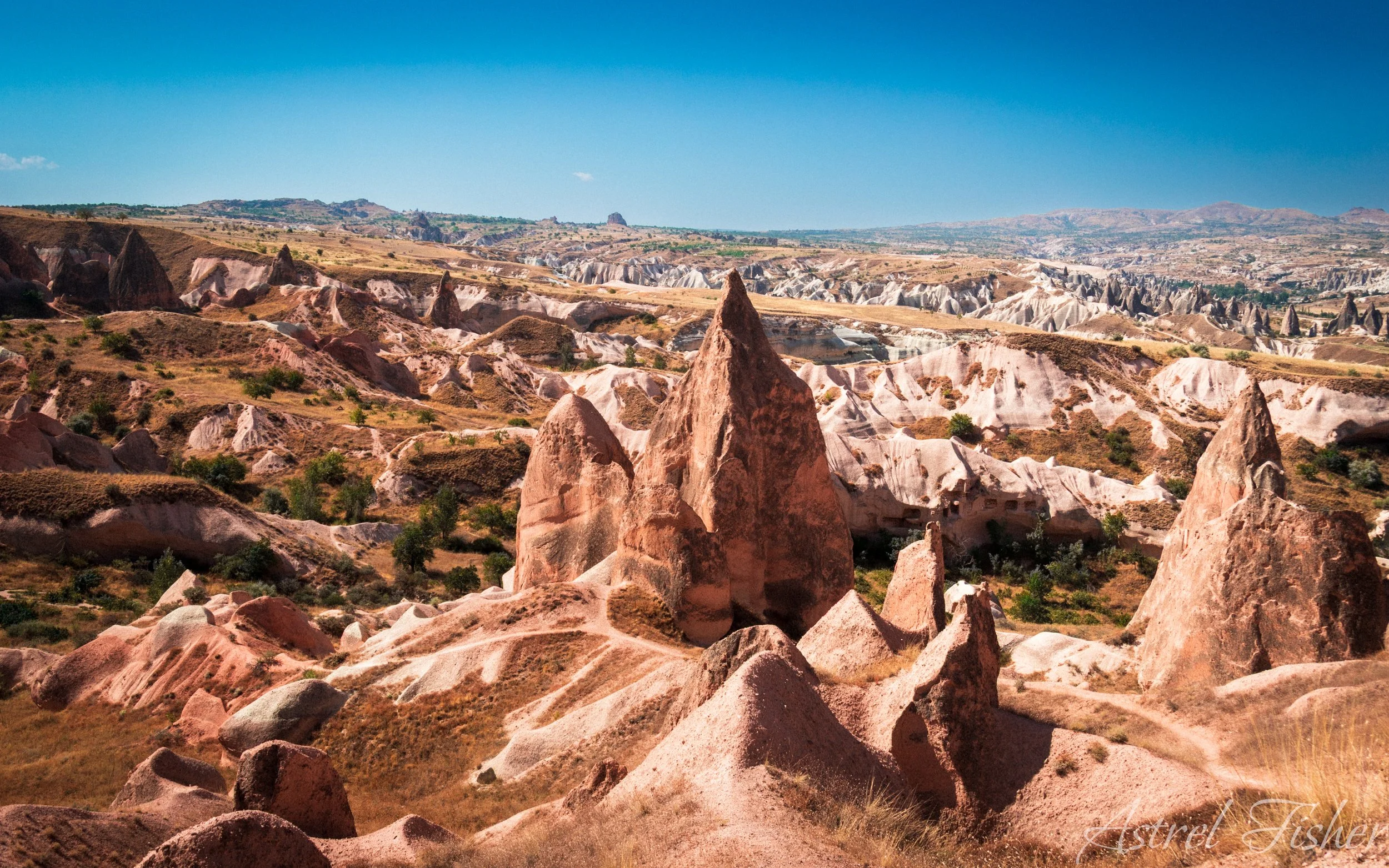 A panoramic view of the unique rock formations and landscape of Cappadocia, Turkey, with tall, cone-shaped rocks, rolling hills, and a clear blue sky.