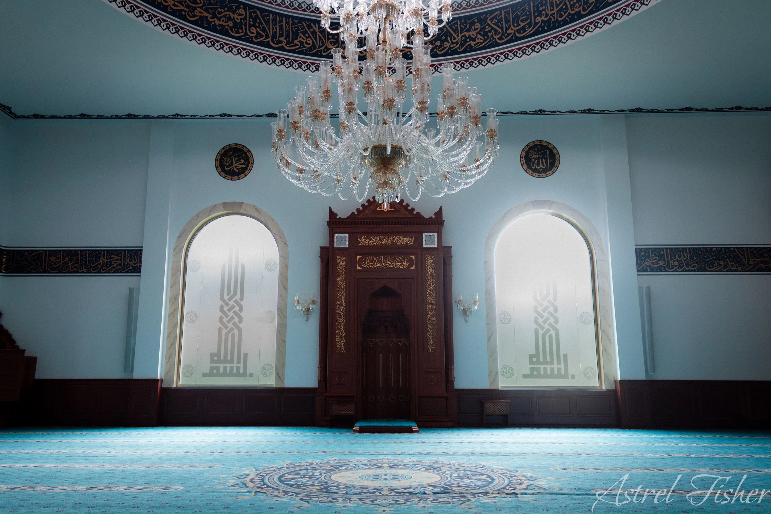 Interior of a mosque with a large crystal chandelier hanging from the ceiling, arched windows allowing natural light, a prayer niche (mihrab) with intricate arabesque designs and calligraphy, and a carpeted floor with a central circular pattern.